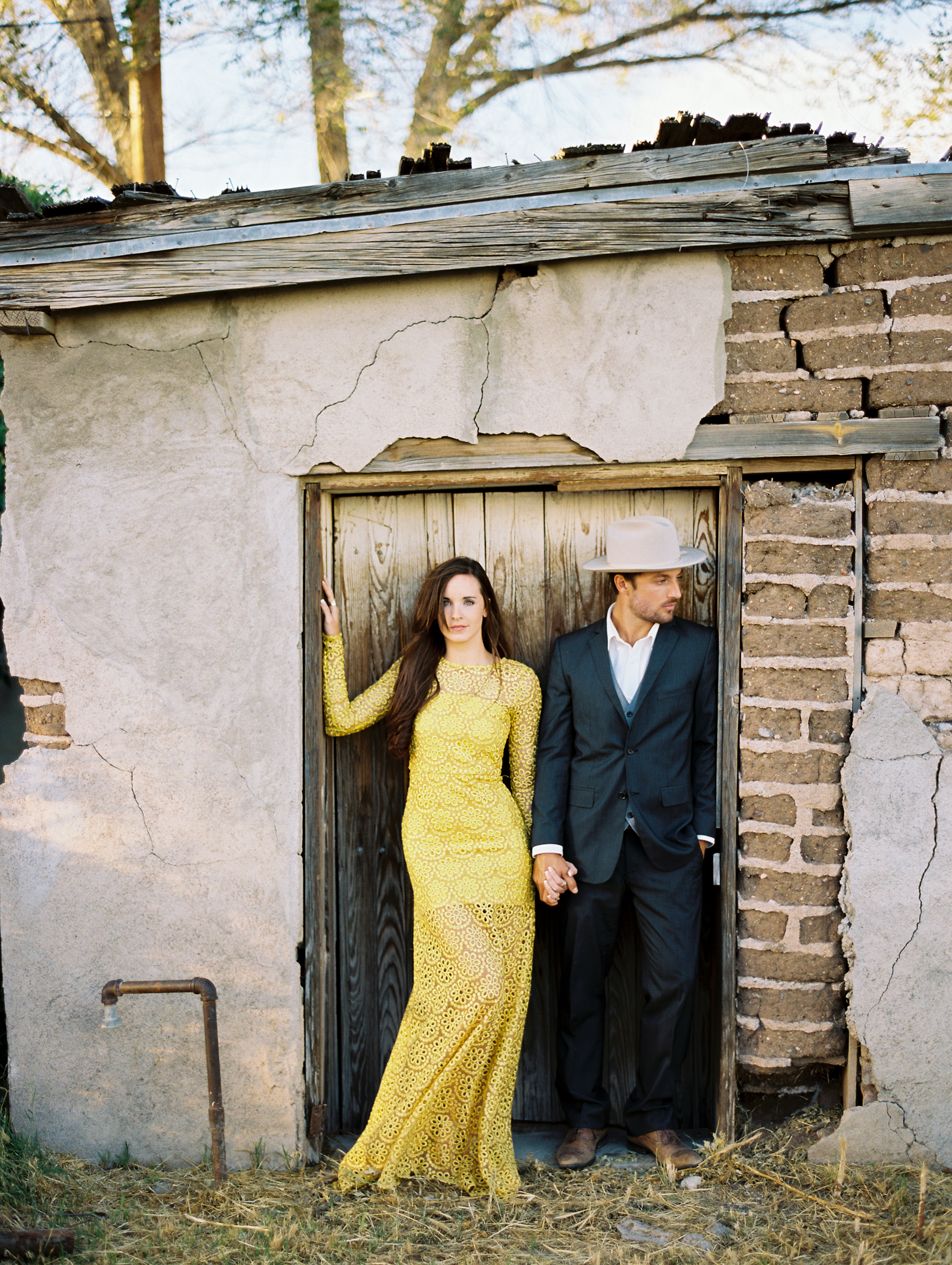 woman in yellow dress holding hands with man in grey suite and cowboy hat standing in wooden doorway engagements in Marfa, Texas.