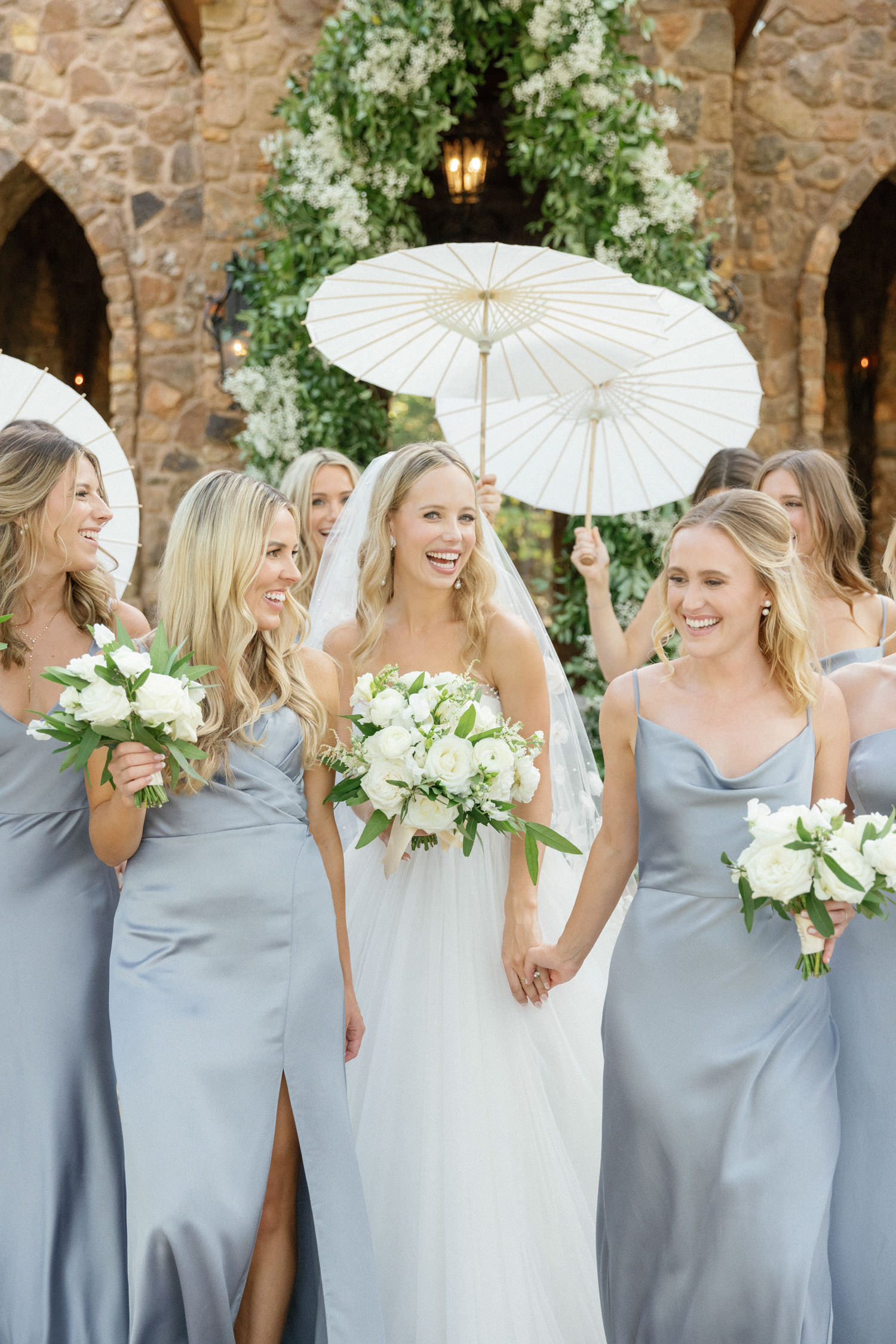 Bride walking with bridesmaids in blue dresses holding umbrellas and laughing texas tented ranch wedding