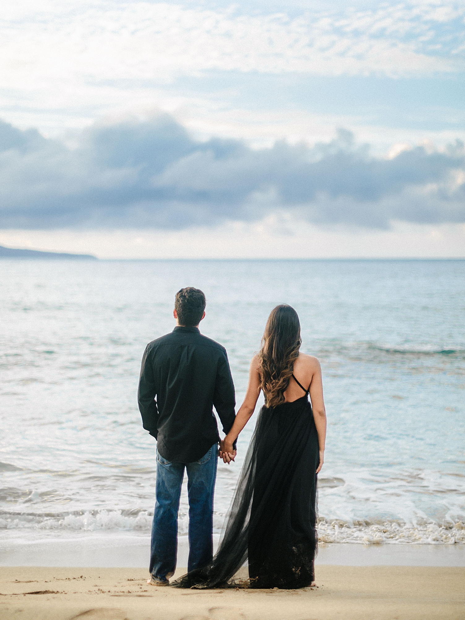 couple in black dress holding hands looking out to ocean on maui beach engagement