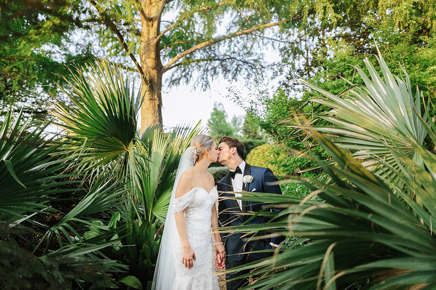 Groom in blue suit kissing bride in white dress and veil standing in green palms destination wedding locations