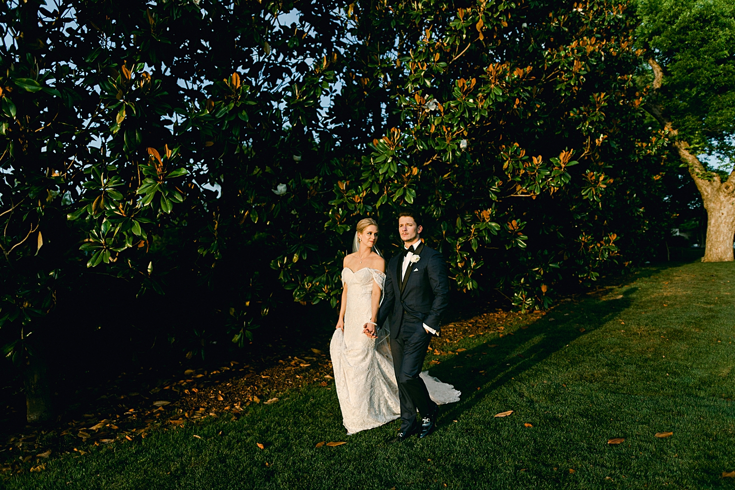 Bride in white wedding dress and veil and Groom in blue tuxedo walking and holding hands in front of magnolia trees at Dallas wedding