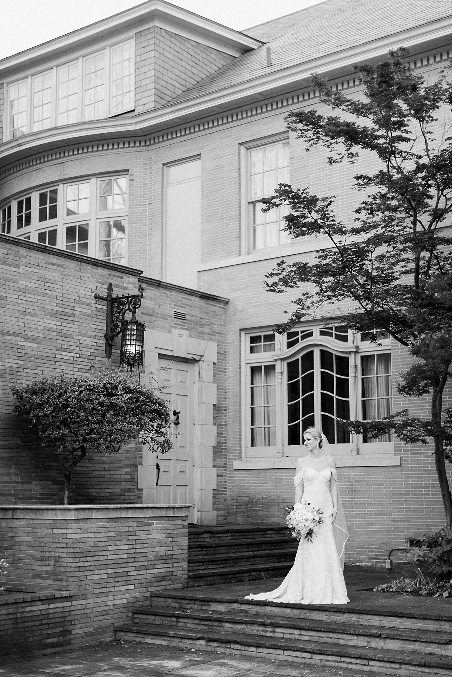 bride in white wedding dress holding floral bouquet and laughing in front of brick home bridal session  black and white