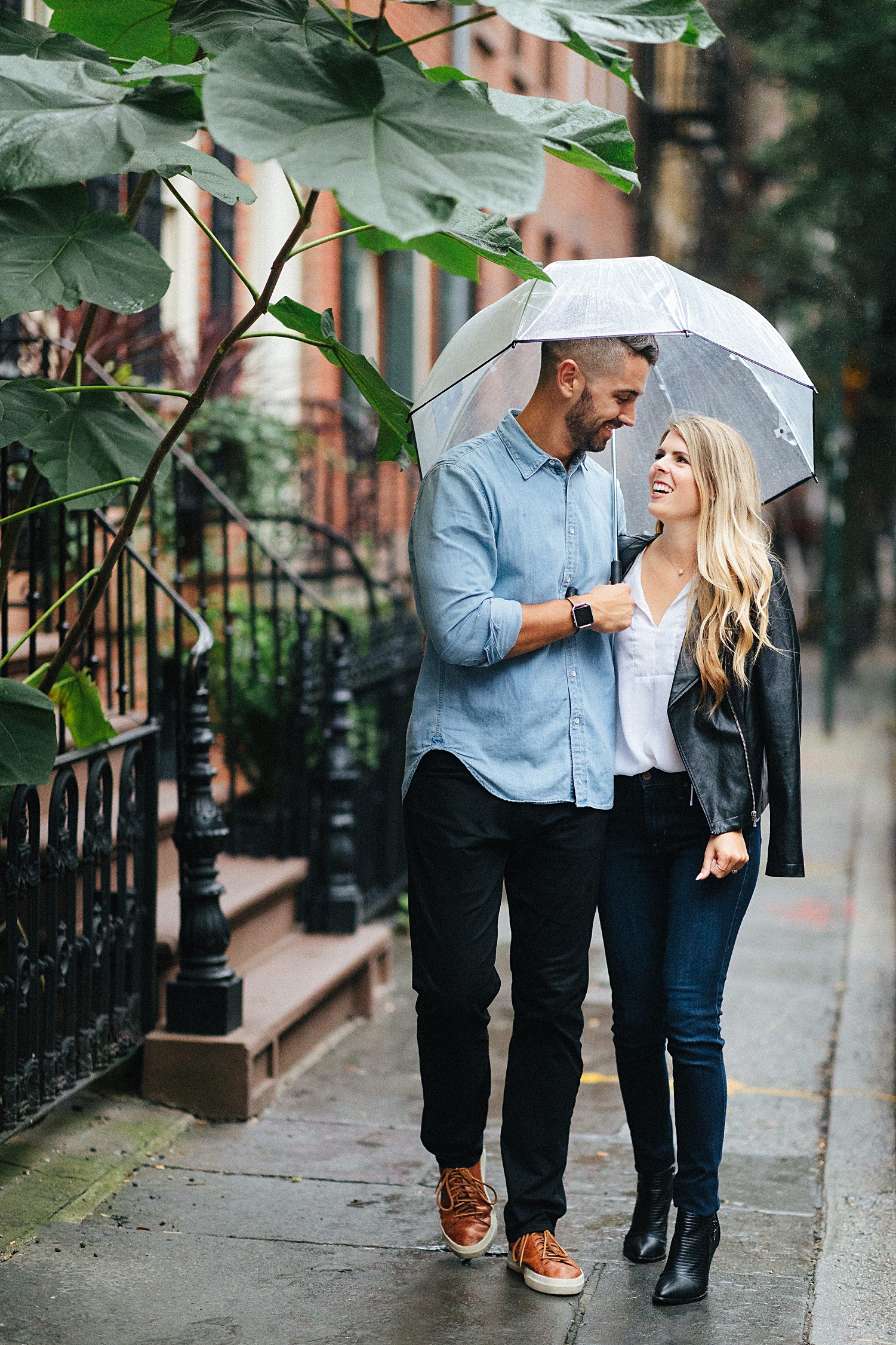 couple walking on sidewalk holding clear umbrella in Greenwich Village engagement