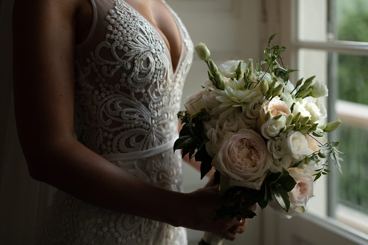 Bride holding floral bouquet in white lace wedding dress by window