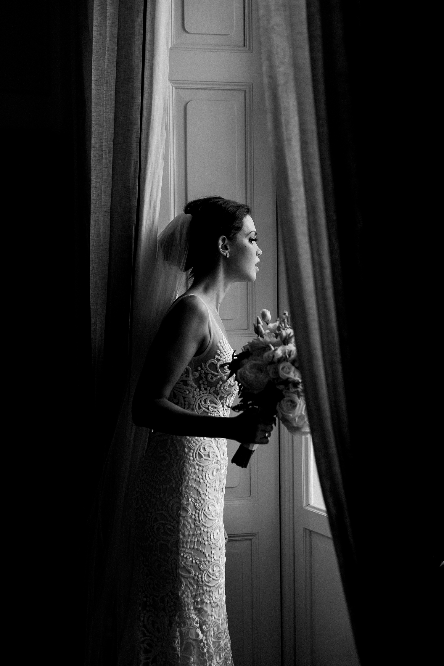 Bride holding floral bouquet in white lace wedding dress by window black and white