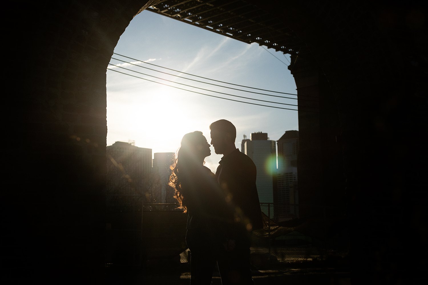 couple standing in archway overlooking downtown manhattan sunset light flare sky
