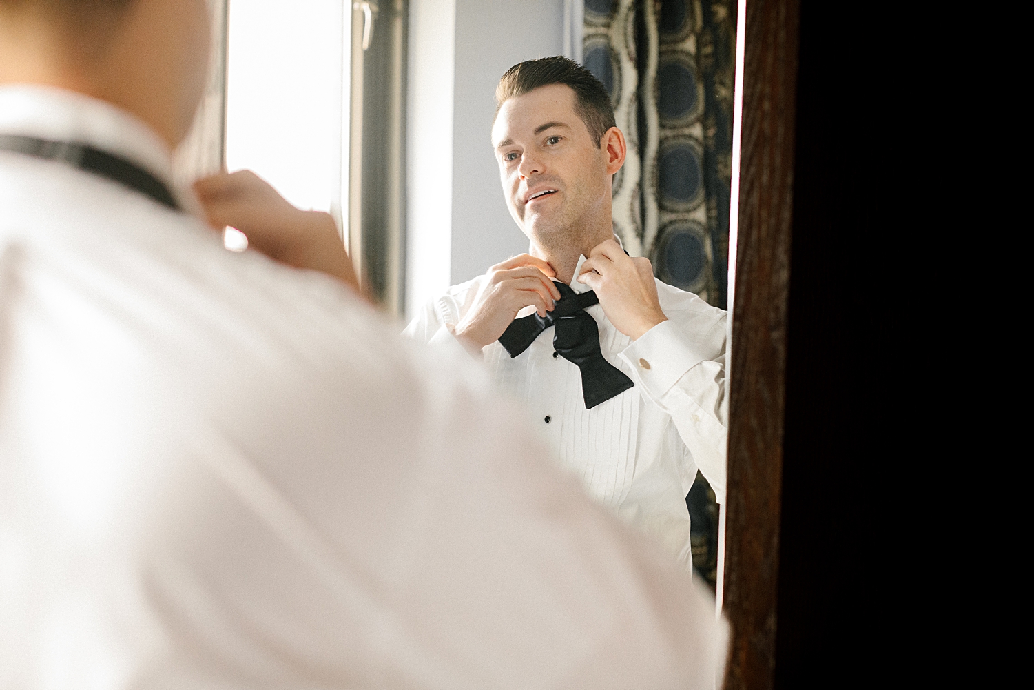 man tying black bow tie looking in mirror