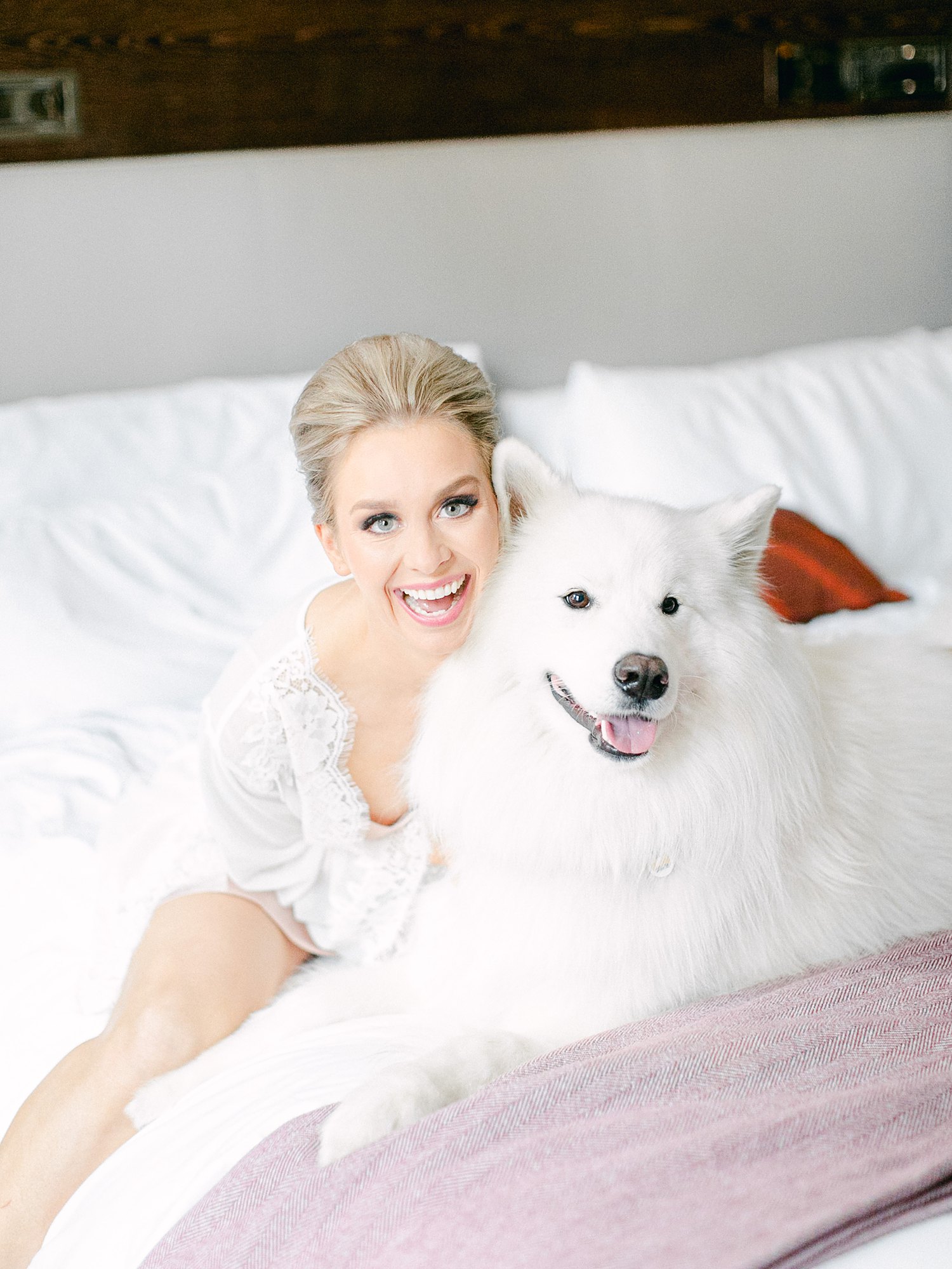 girl in white robe on bed with white fluffy dog smiling at Dallas Arboretum Wedding