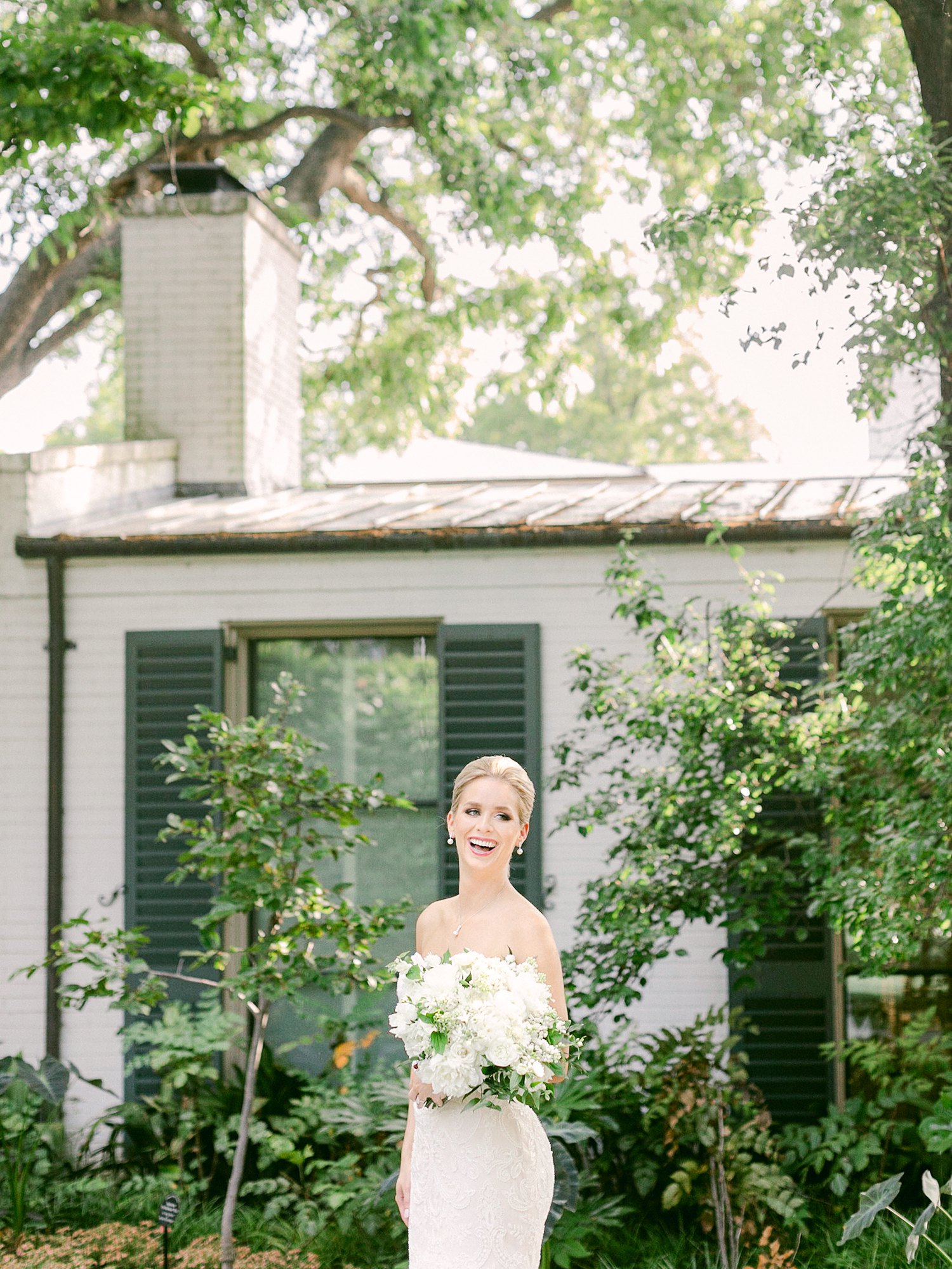 Bride holding white bouquet laughing in front of building with greenery at Dallas Arboretum Wedding