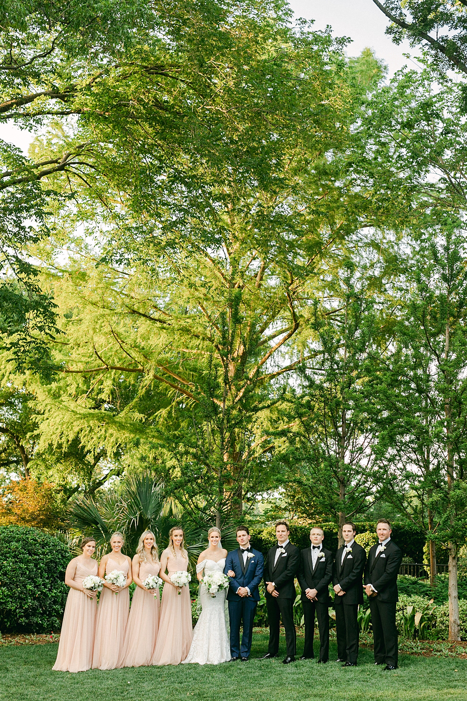 Bride in white wedding dress and veil and Groom in blue tuxedo posing with four bridesmaids in peach dresses and groomsmen in black tuxedos for in green garden