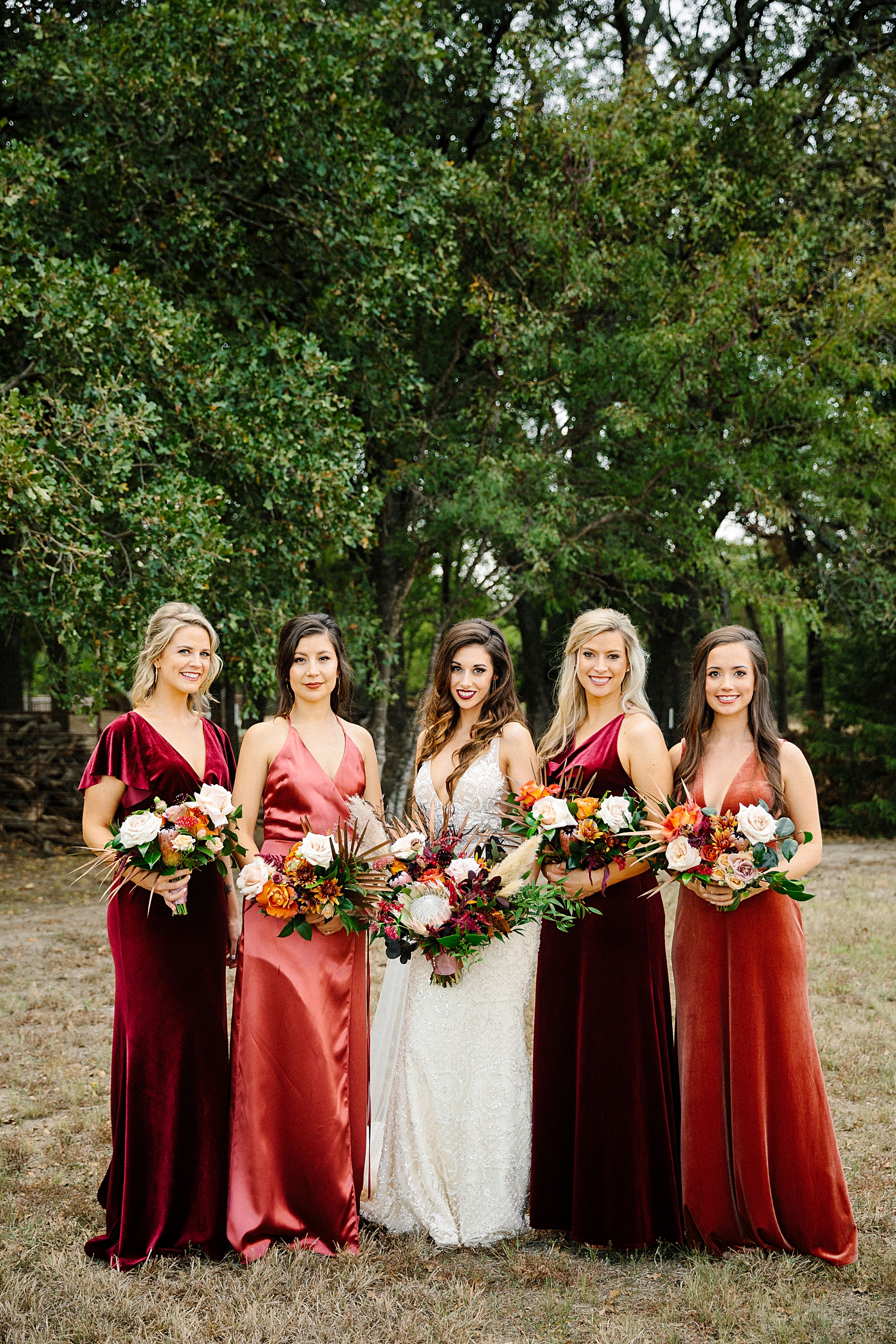 bride and bridesmaids in red dresses holding floral bouquets  standing in front of green trees