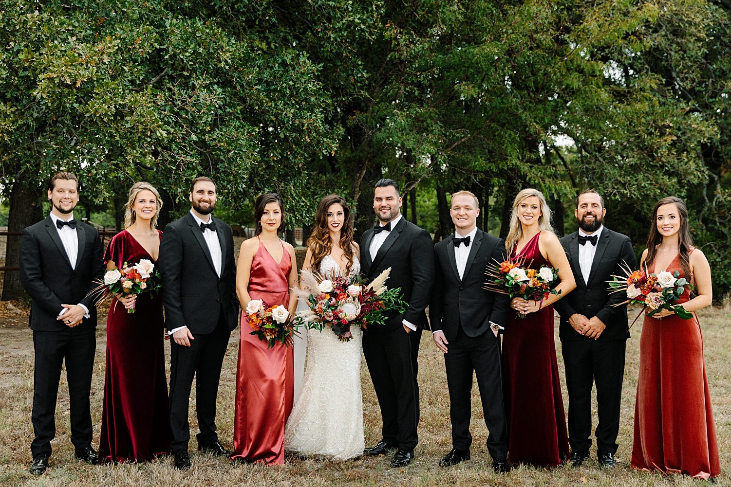 bride groomsmen and bridesmaids in red dresses holding floral bouquets standing in front of green trees Emerson Venue Wedding