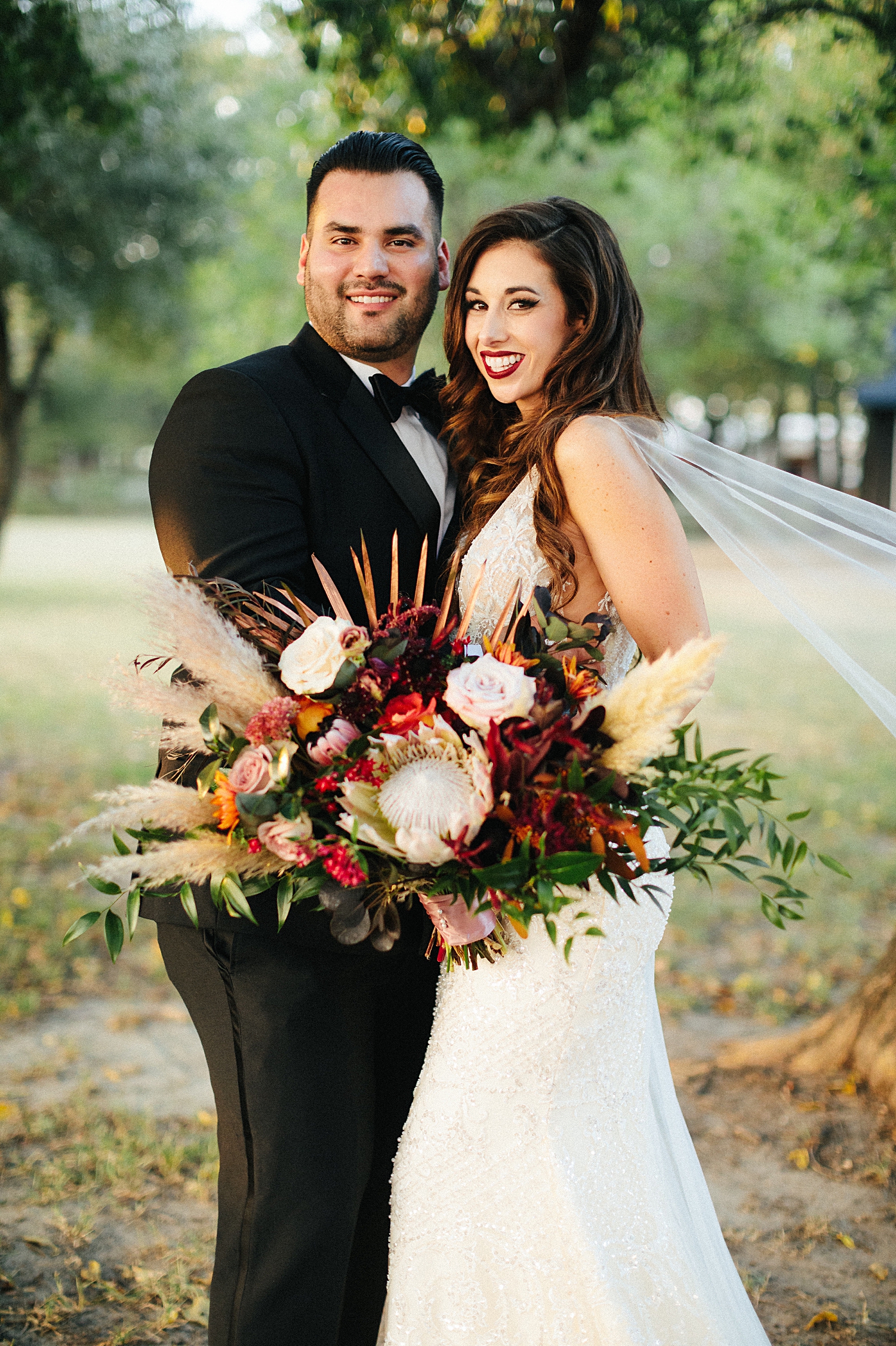 bride in white dress with cape veil embracing groom in black tuxedo holding large bridal bouquet smiling at Emerson Venue Wedding