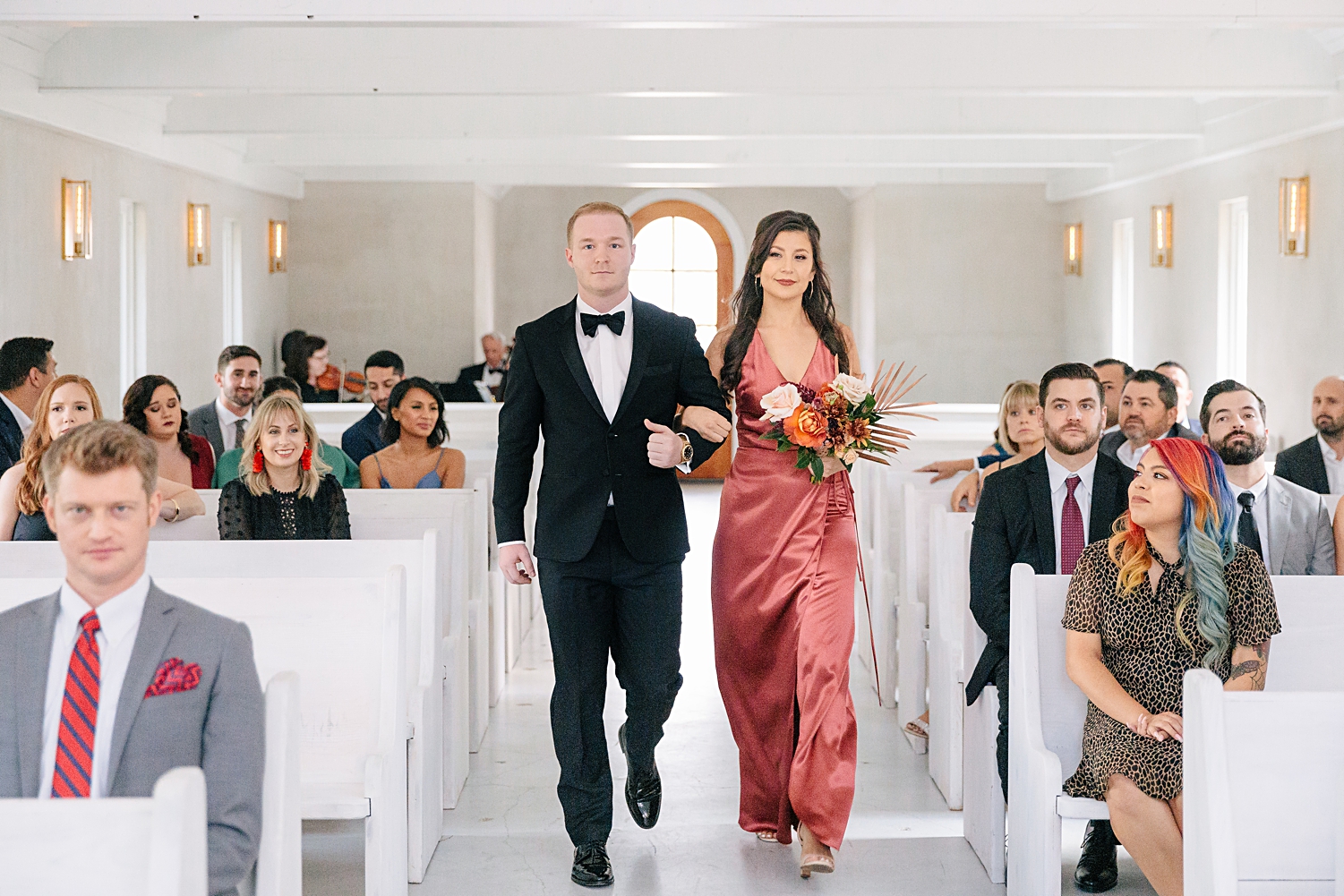 groomsman and bridesmaid in rose gold dress walking down aisle of white chapel wedding