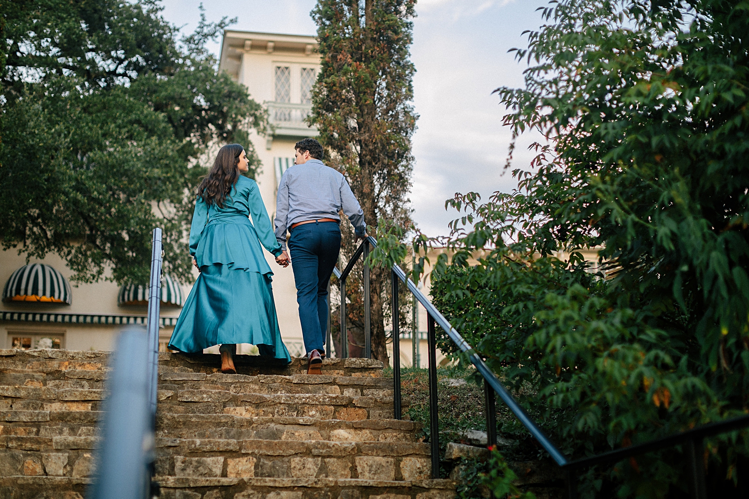 woman in blue dress walking up stone stairs with man in front of Spanish mansion