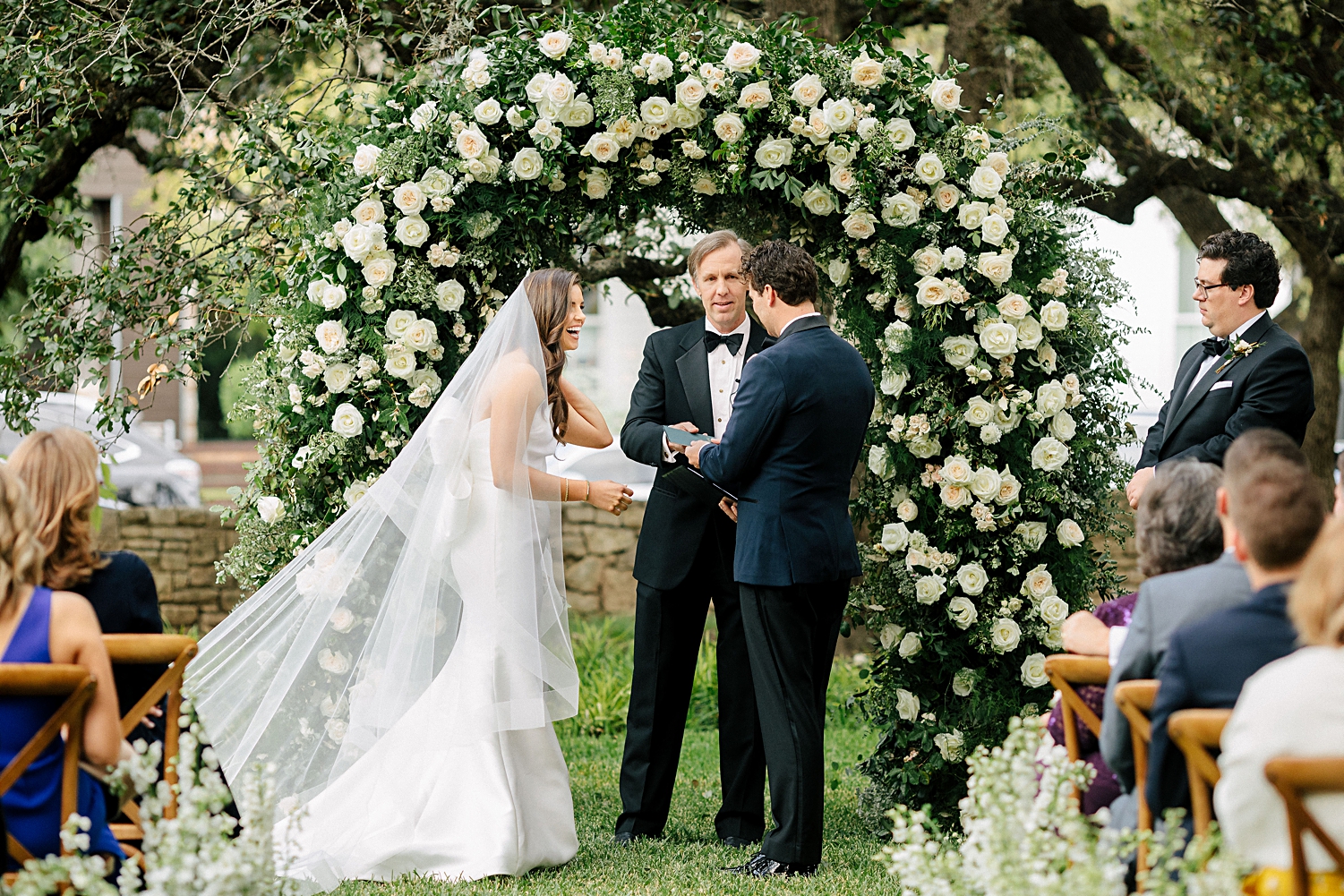 Bride laughing with groom in navy tuxedo at ceremony altar in front of white floral arch