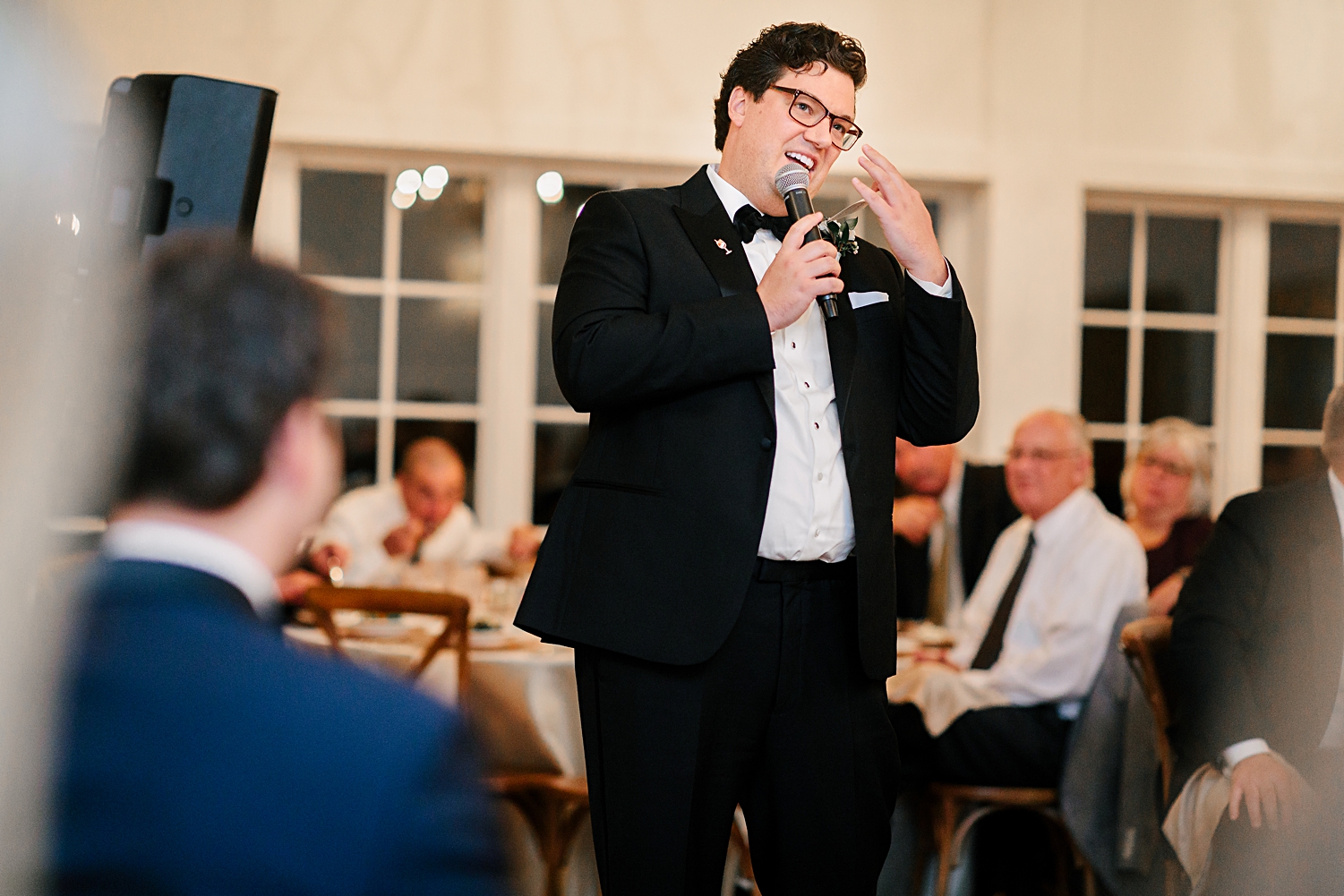 groomsman in black tuxedo giving speech at wedding reception