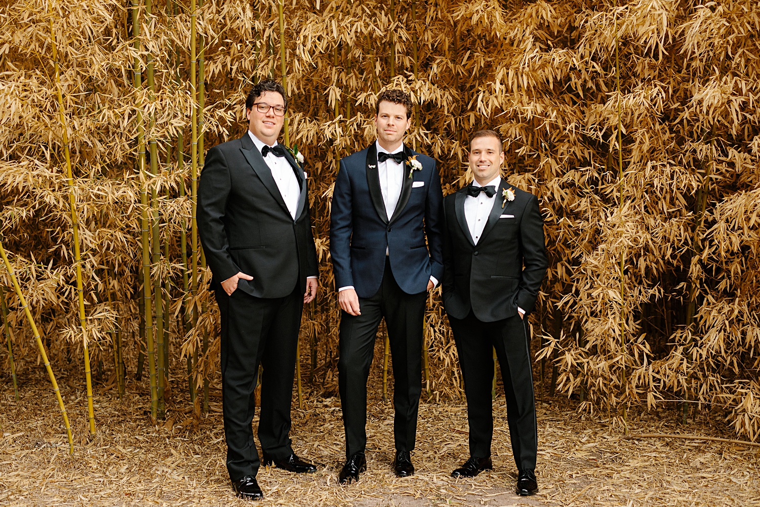 Groom in blue tuxedo standing with two groomsmen in black in front of golden plants at Mattie's At Green Pastures Austin wedding