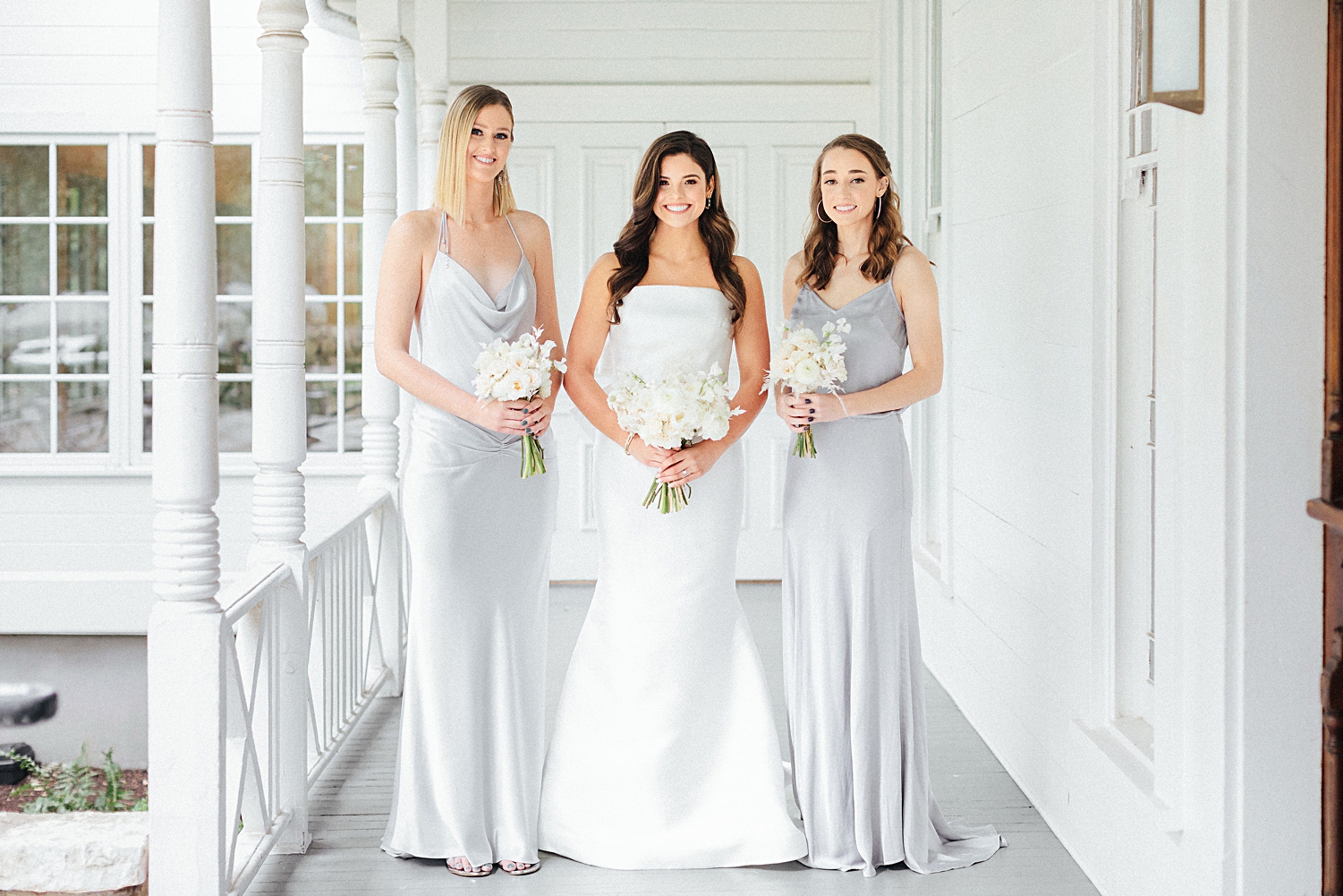 smiling bride in strapless white wedding dress standing with two bridesmaids in silver dresses bouquet