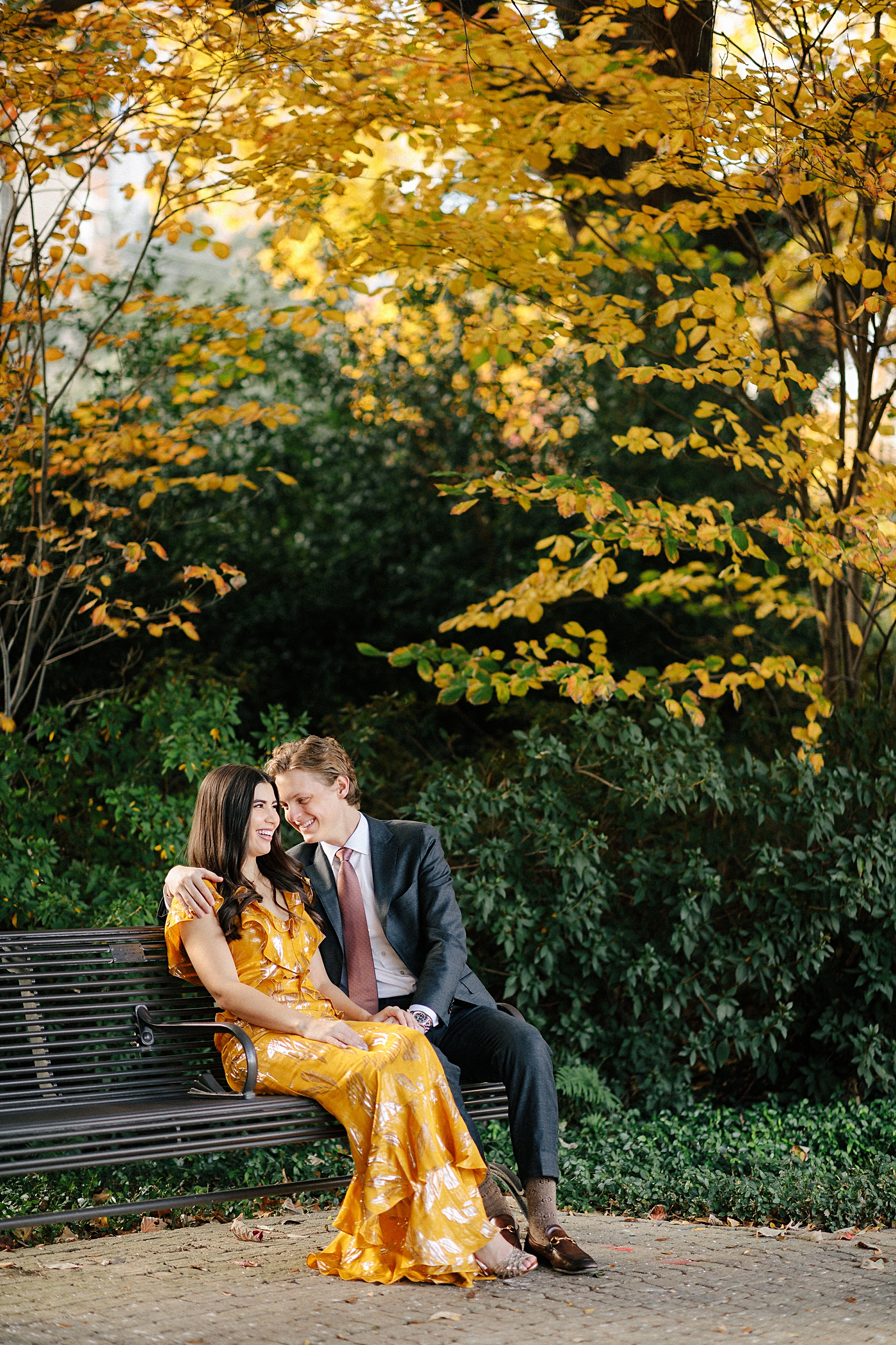 man in grey suit smiling with woman in yellow dress while sitting on park bench