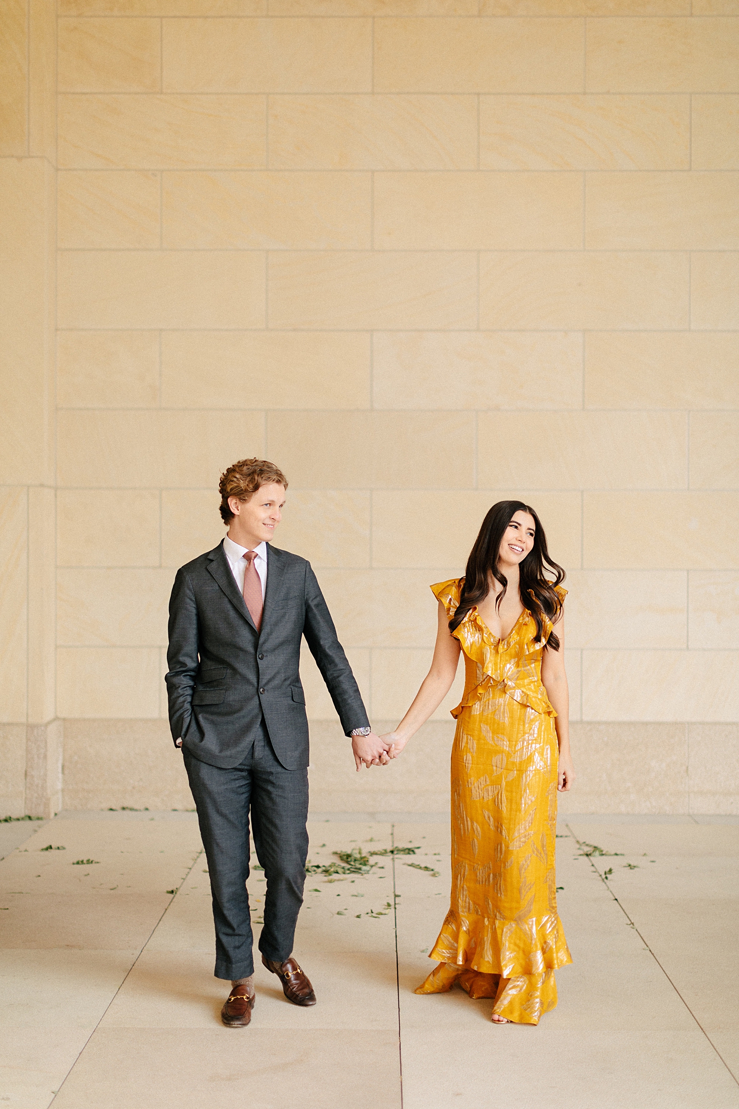 woman in yellow dress holding hands with man in suit in front of stone wall engagement smiling