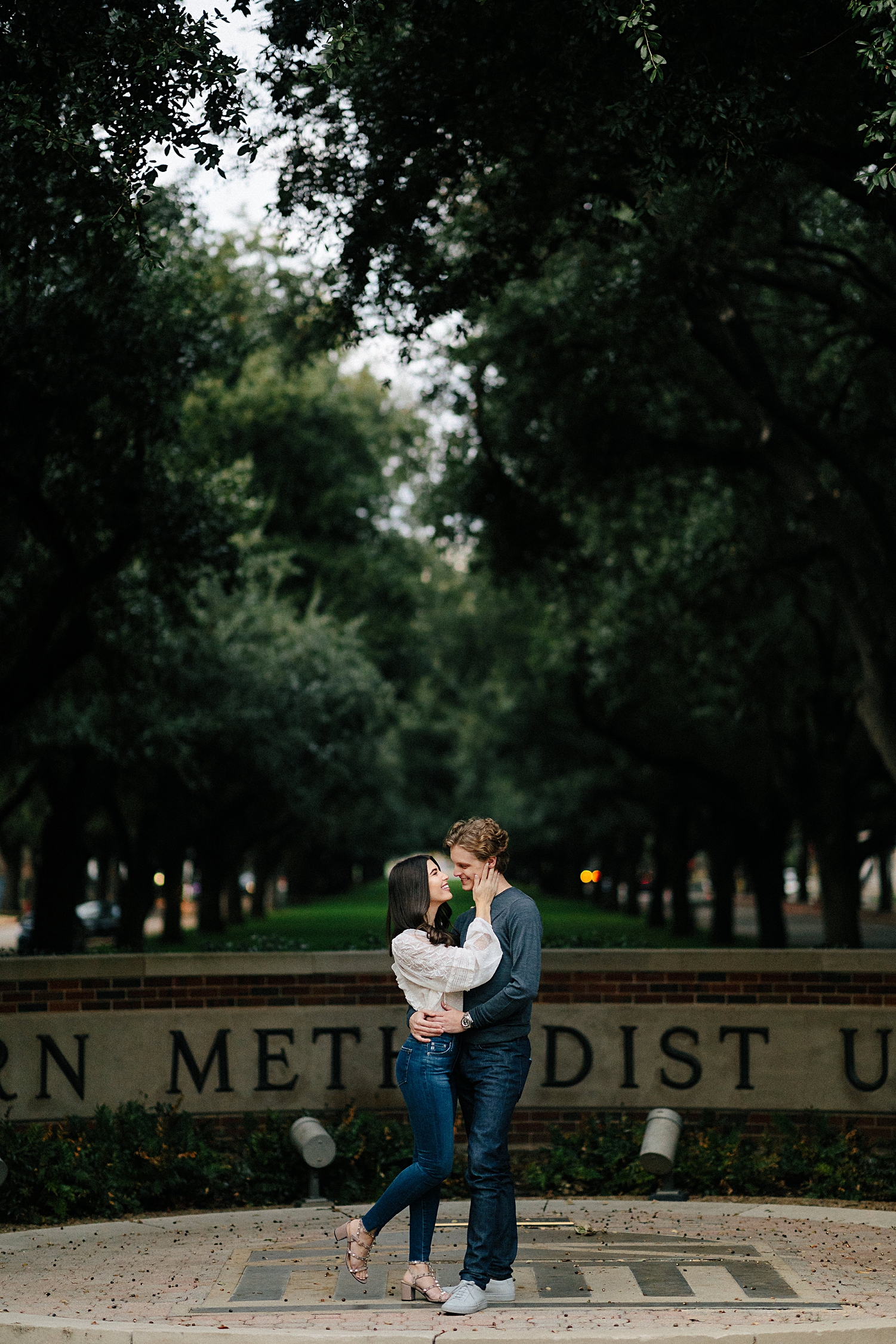 Couple hugging and smiling at each other during engagement in front of Southern Methodist University SMU campus entrance sign