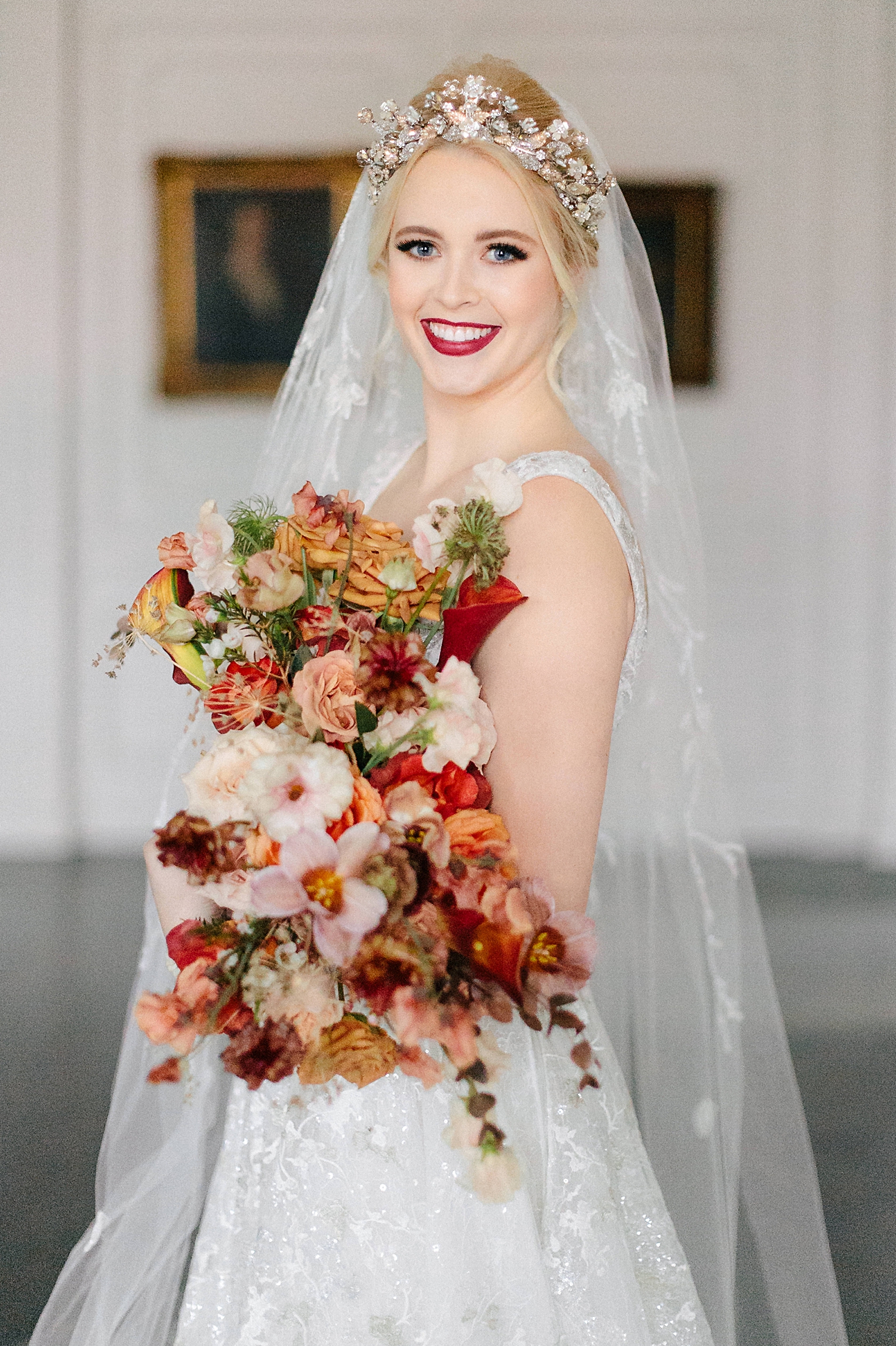 smiling bride in shimmering white wedding dress, gold crown, veil standing in white room holding orange and red bridal bouquet at the Mason Dallas