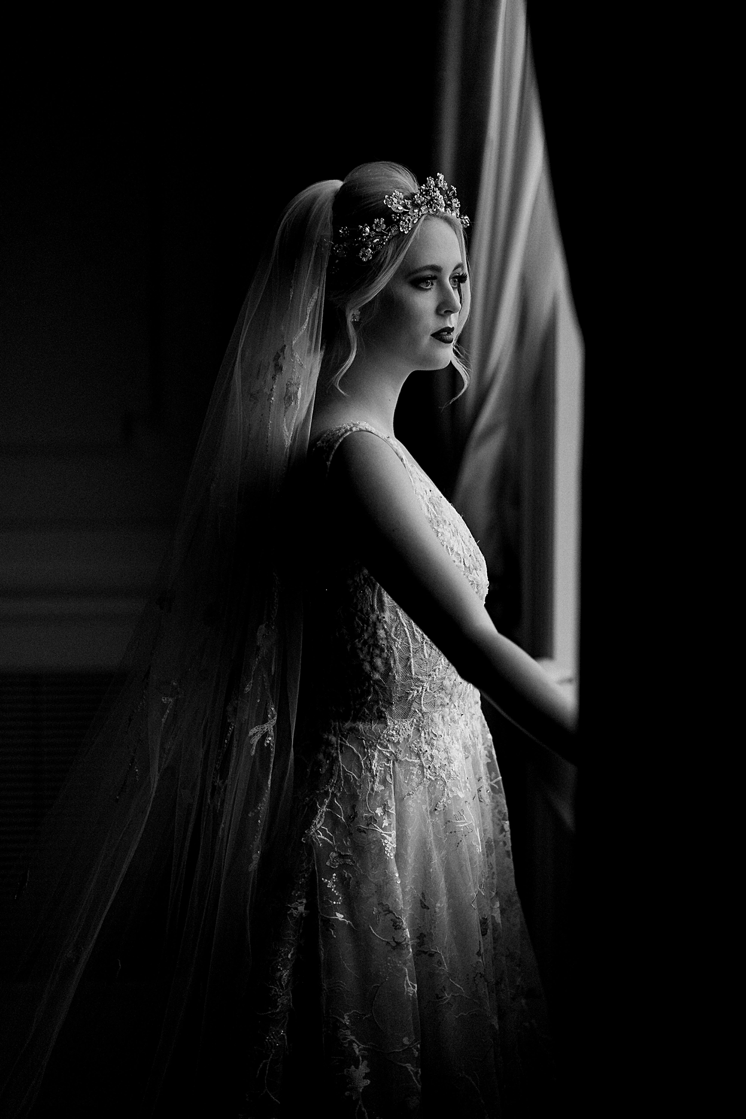bride in wedding dress, crown, veil standing by window in dark room