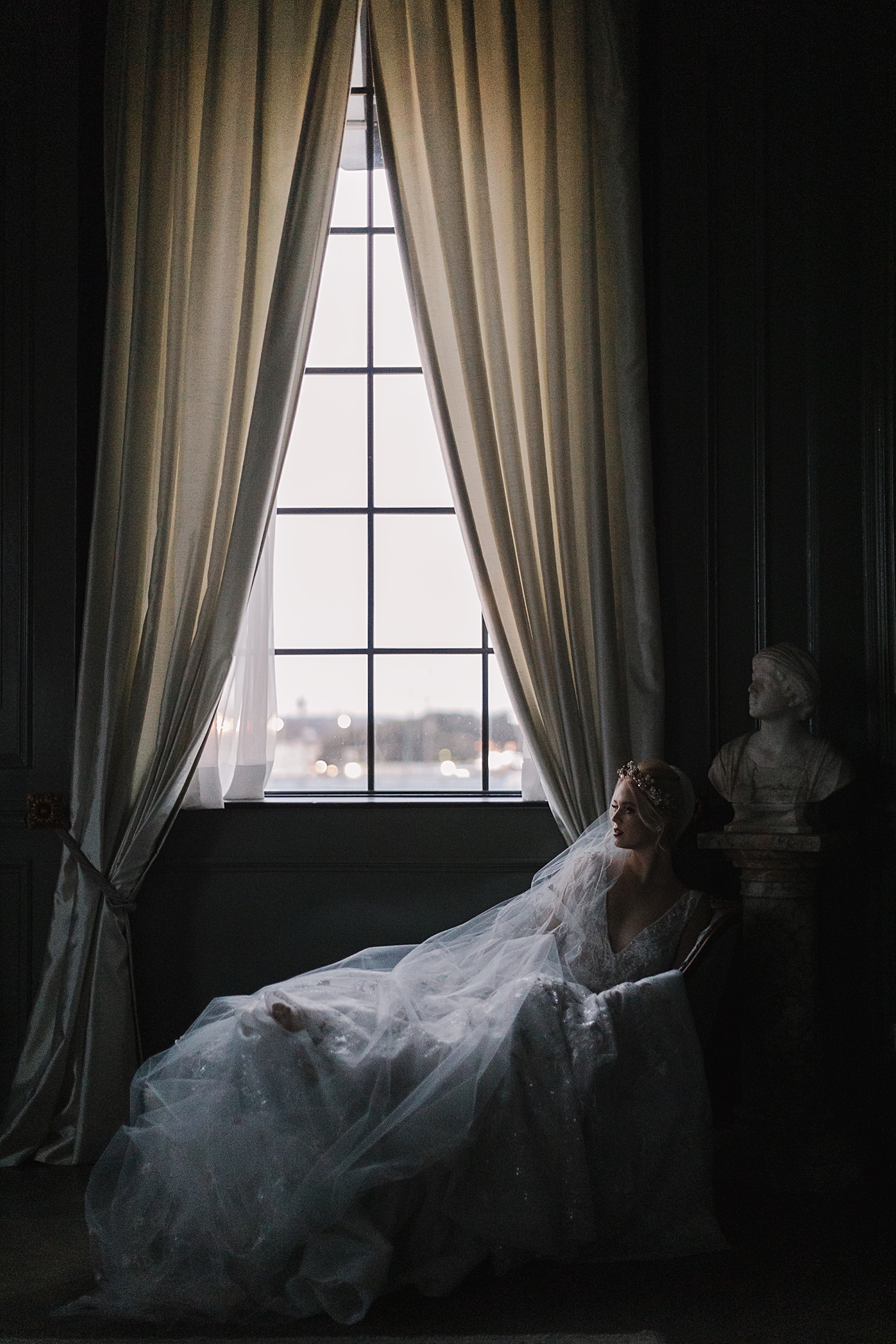 bride in wedding dress, crown, veil laying on duvet by window in dark room The Mason Dallas