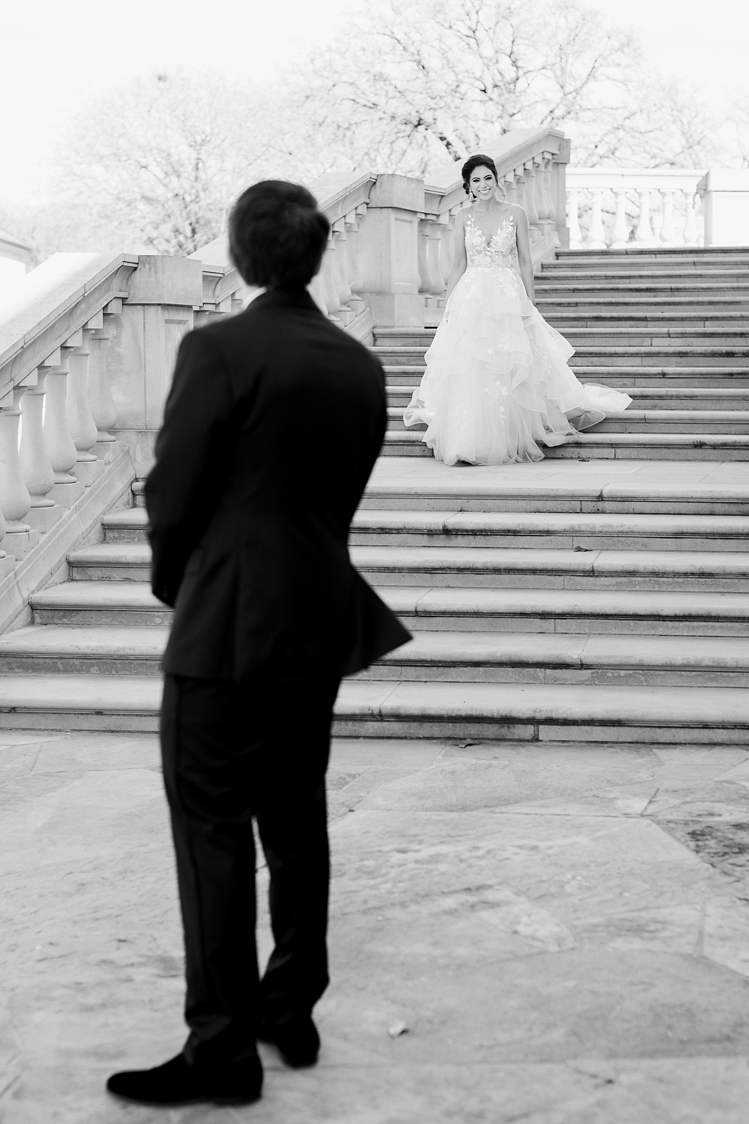 man in black tuxedo waiting for bride in white gown to come down concrete stairs on french estate wedding day black and white
