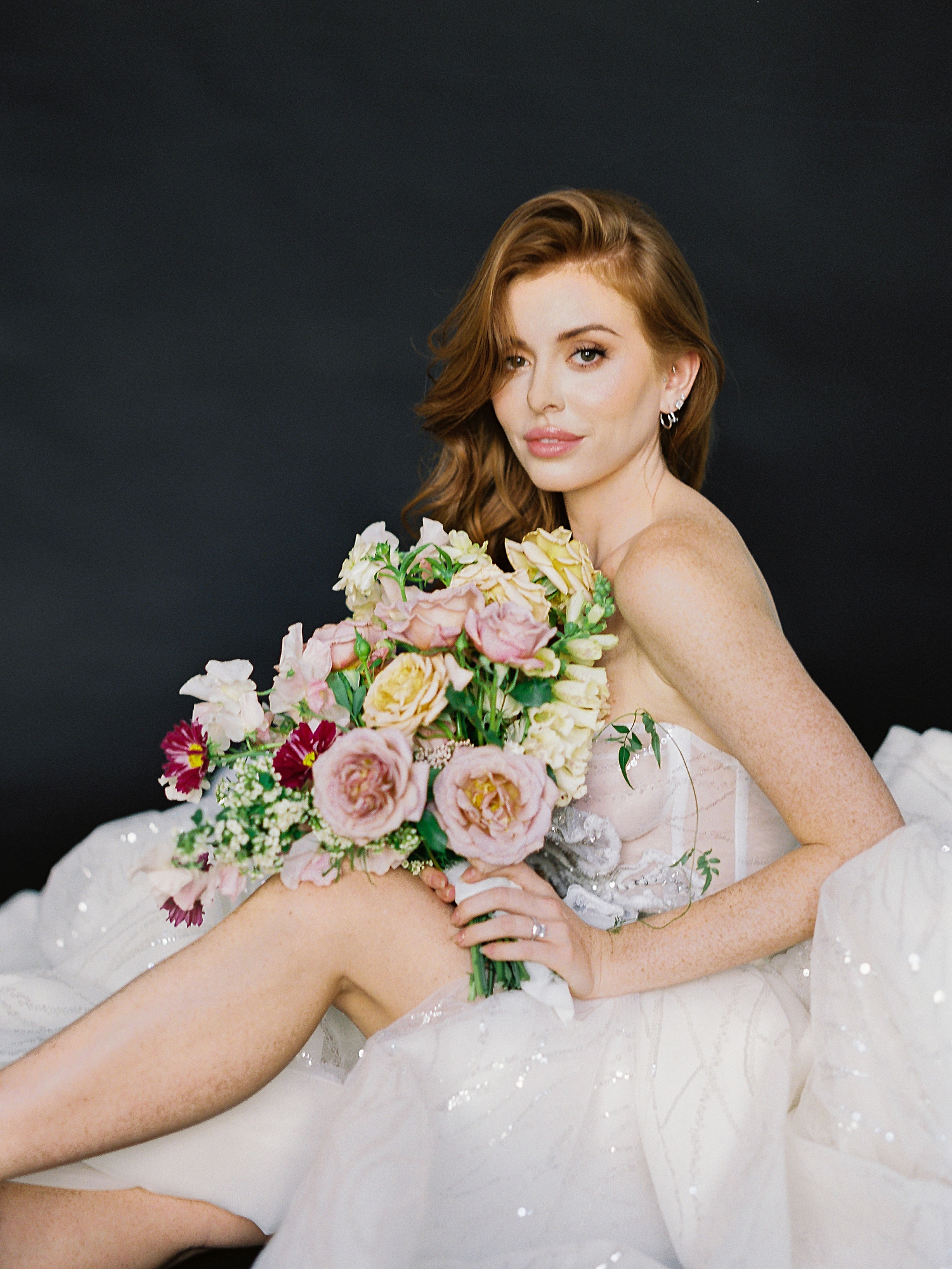red headed girl in sparkling white wedding gown sitting with colorful floral bouquet