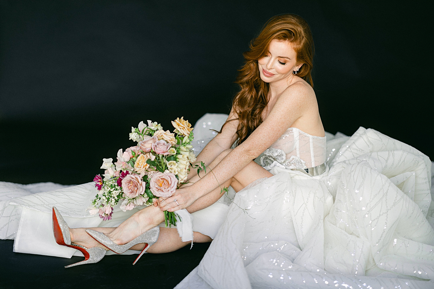 red headed girl in sparkling white wedding and high heels gown sitting with colorful floral bouquet