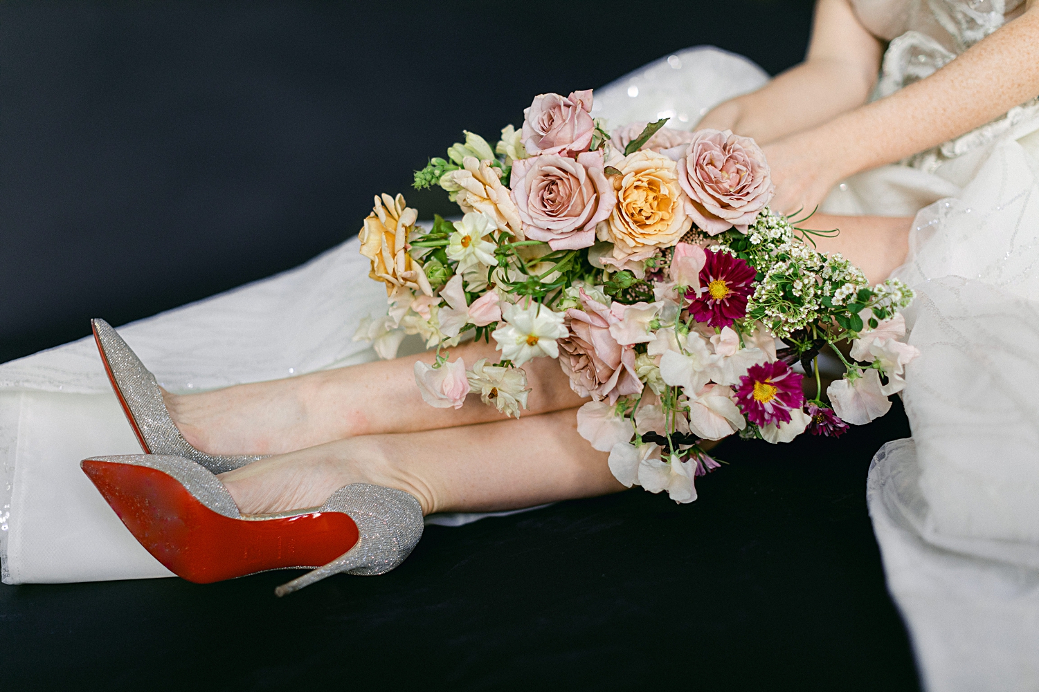 Bride in sparkling white wedding and high heels gown sitting with colorful floral bouquet