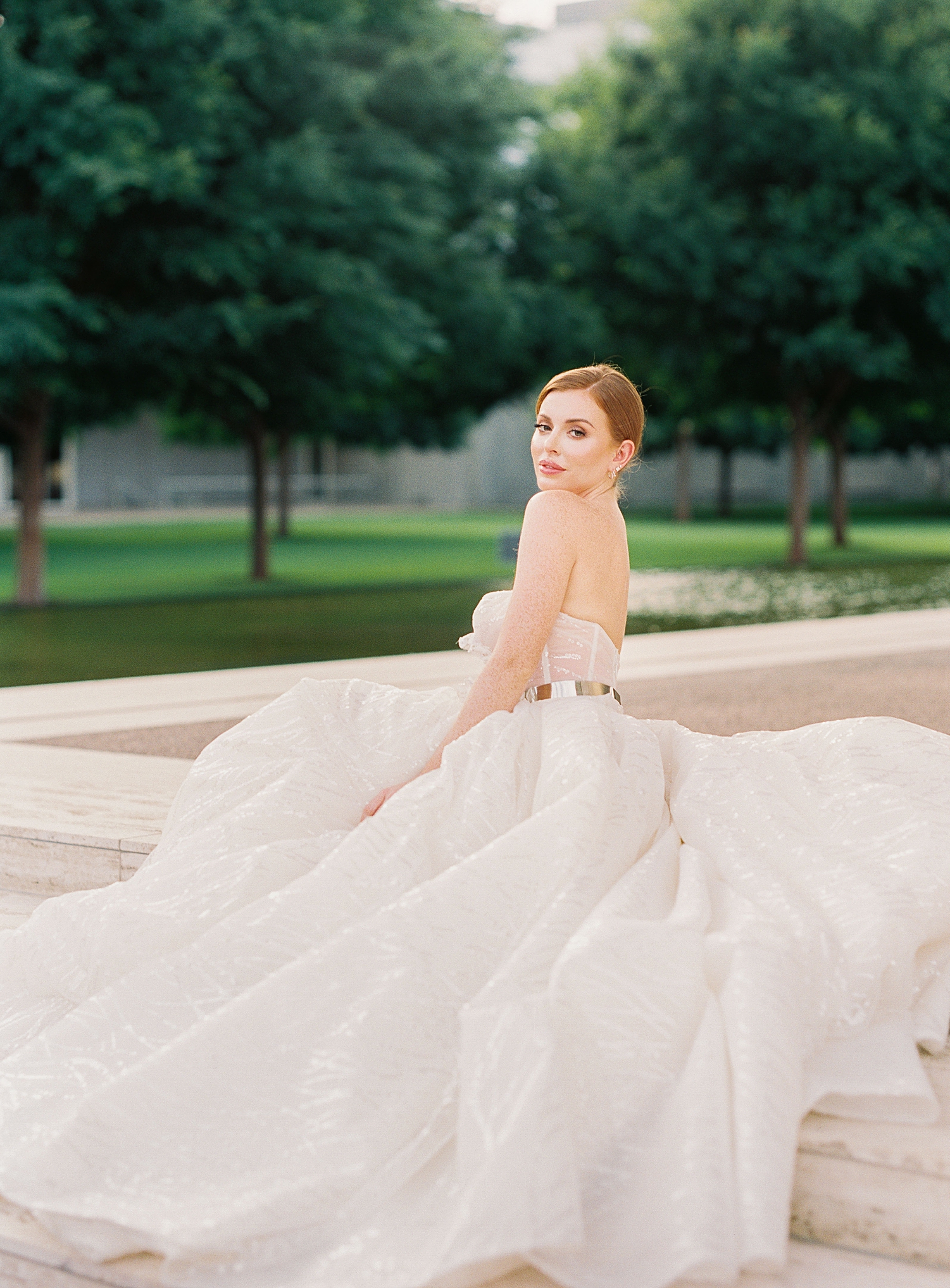 red headed bride in sparkling white Galia Lahav bridal gown sitting in front of green trees