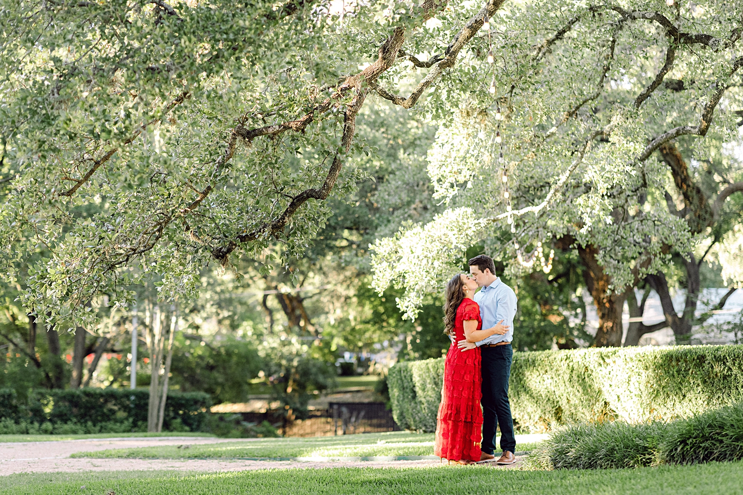 Man and woman in red dress embracing under tree in green garden Austin engagement at Laguna Gloria