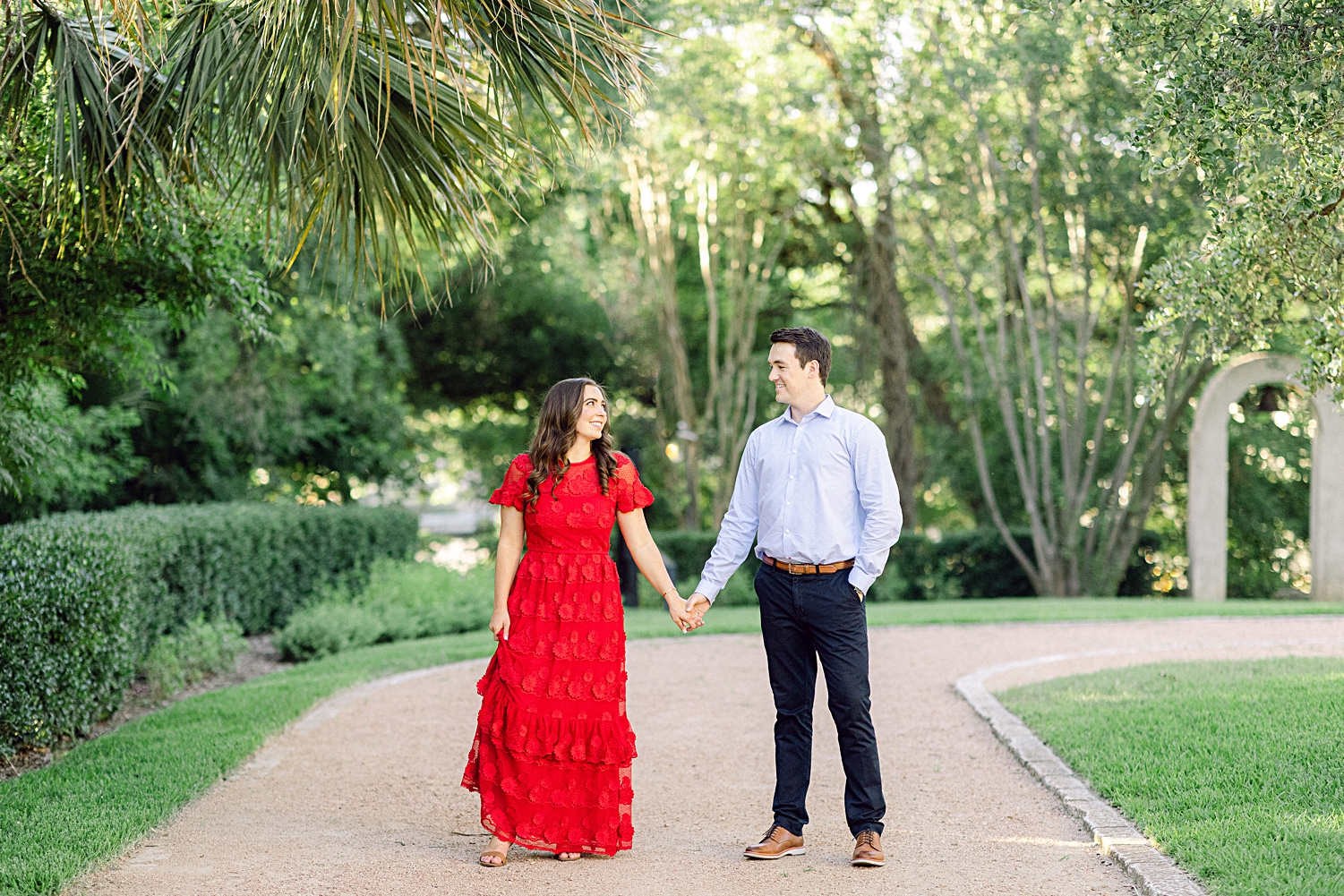 Man and woman in red dress standing holding hands on path in green garden