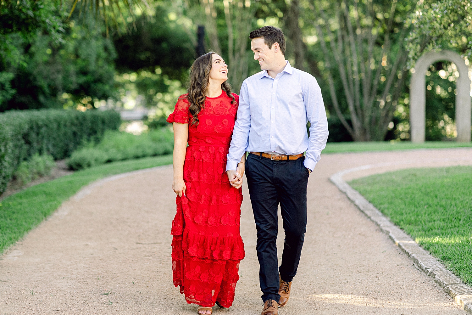 Man and woman in red dress holding hands and walking on path in green garden Austin engagement at Laguna Gloria