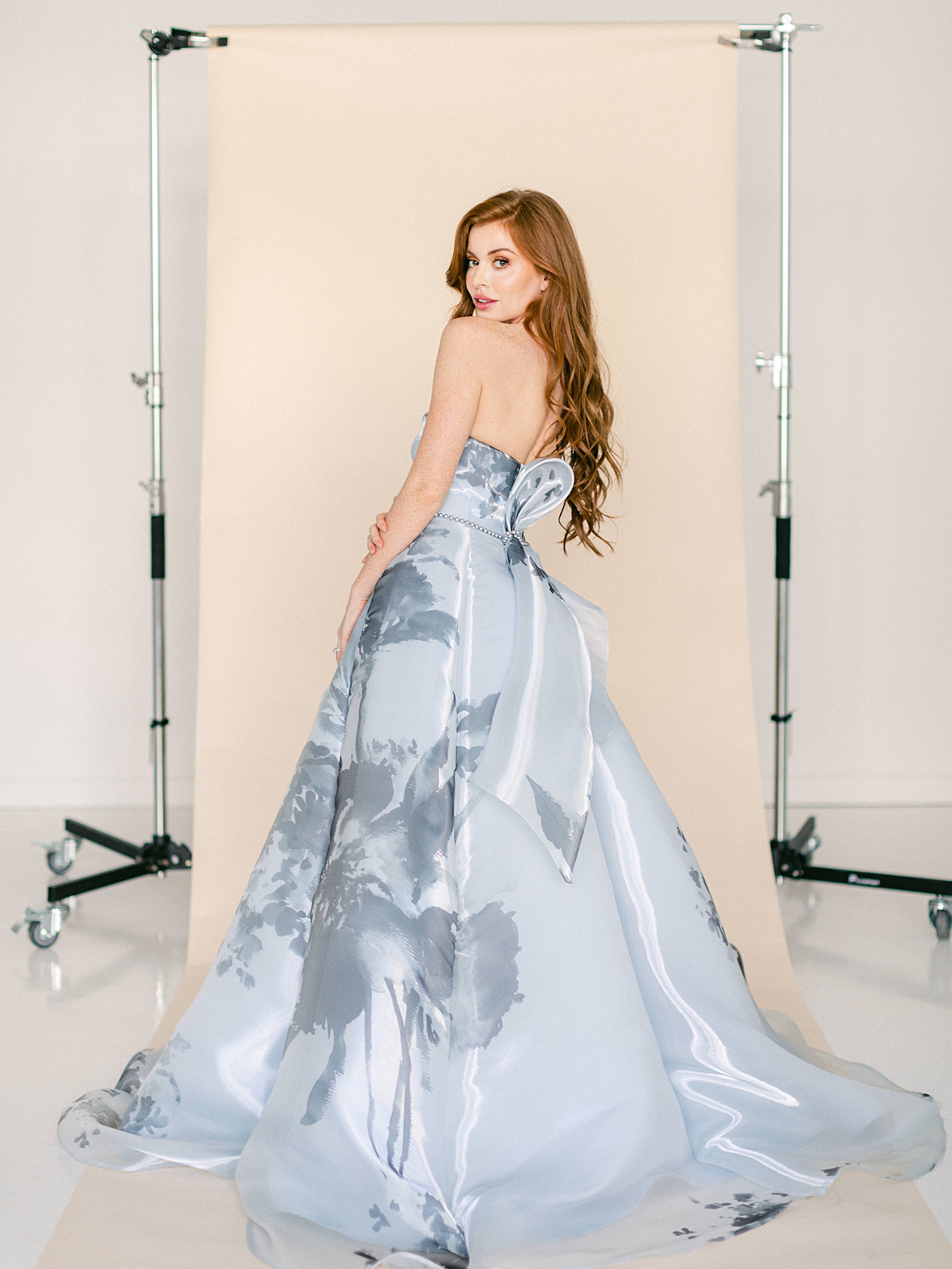 Blue floral ball gown worn by redhead woman looking over shoulder in front of tan backdrop
