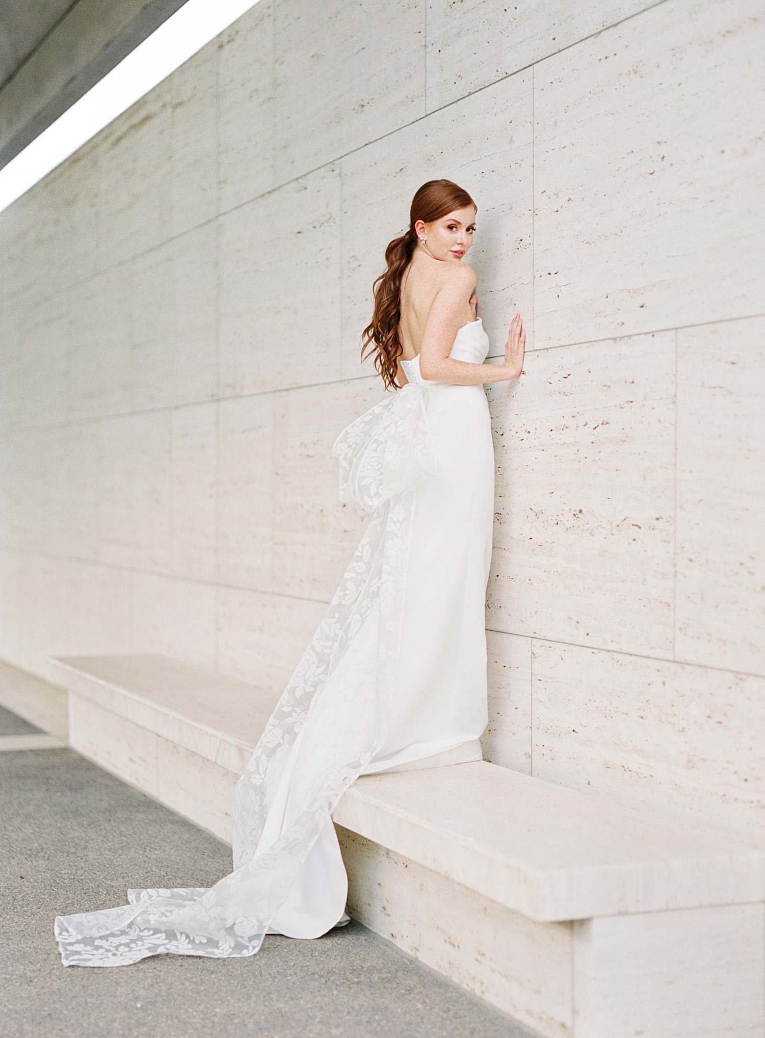 red headed bride standing against concrete wall in white Sareh Nouri bridal gown with lace bow on back