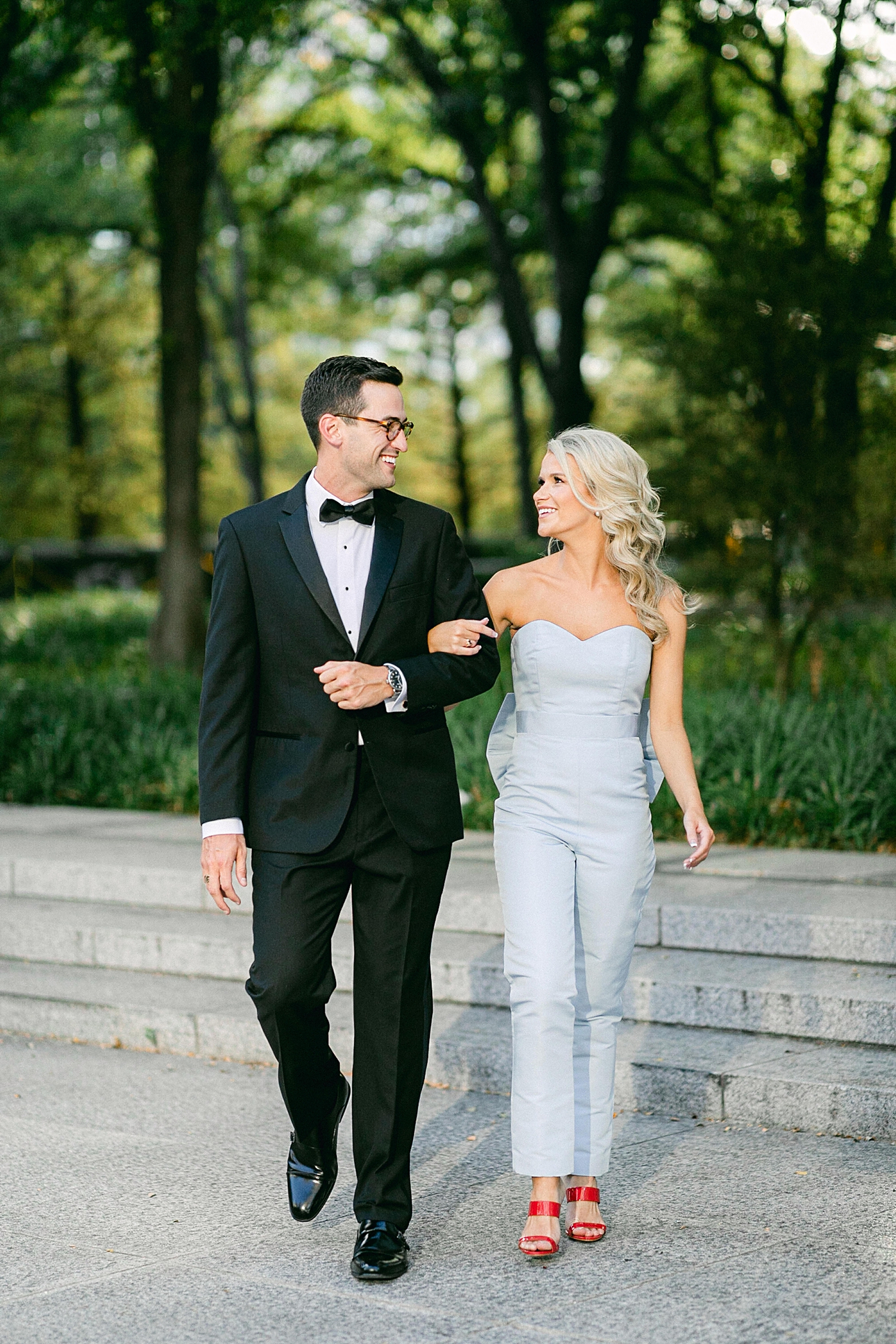 Man in black tuxedo and woman in light blue jumpsuit walking arm in arm by green garden in downtown Dallas engagement