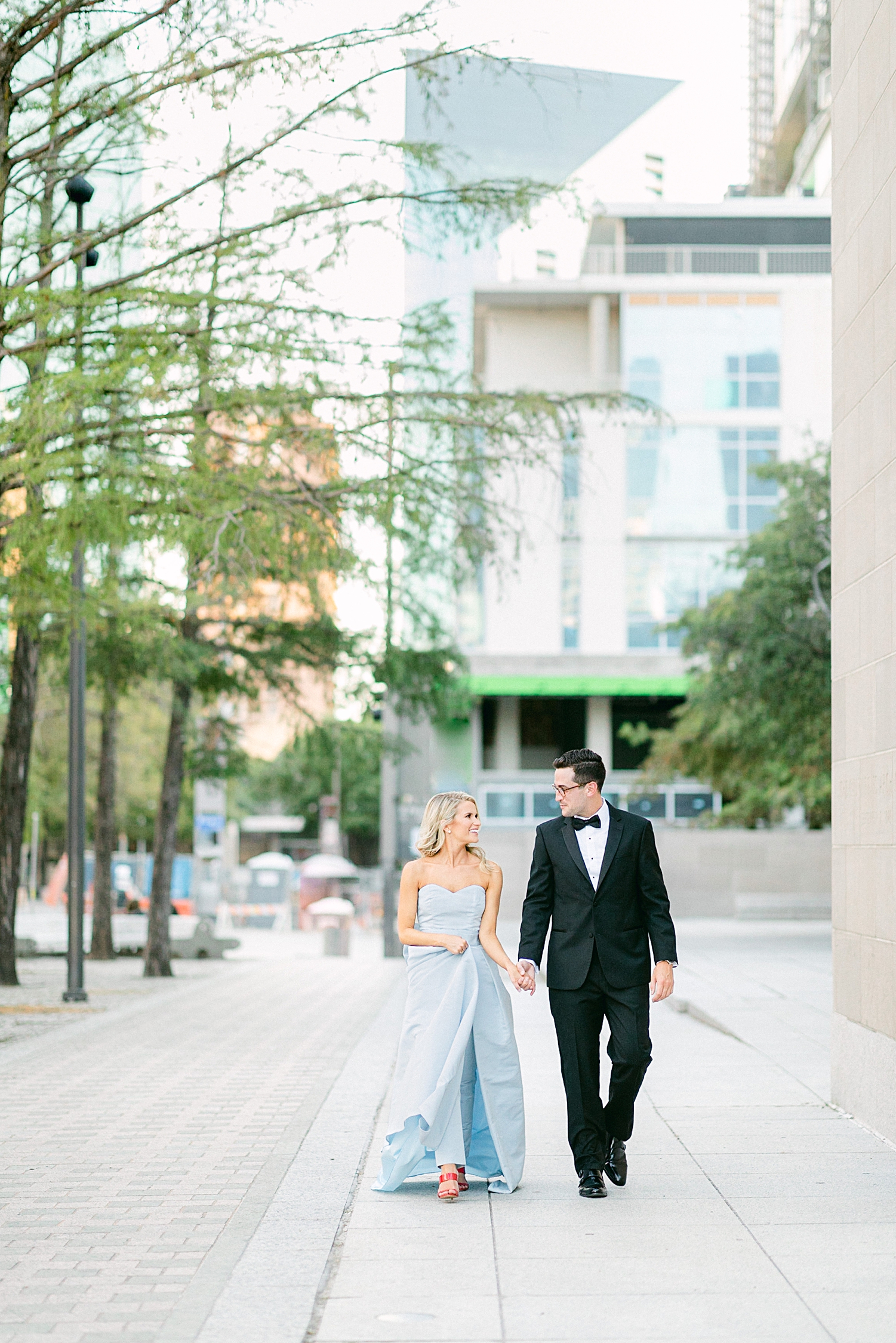 Man in black tuxedo and woman in light blue jumpsuit walking together in downtown Dallas engagement session