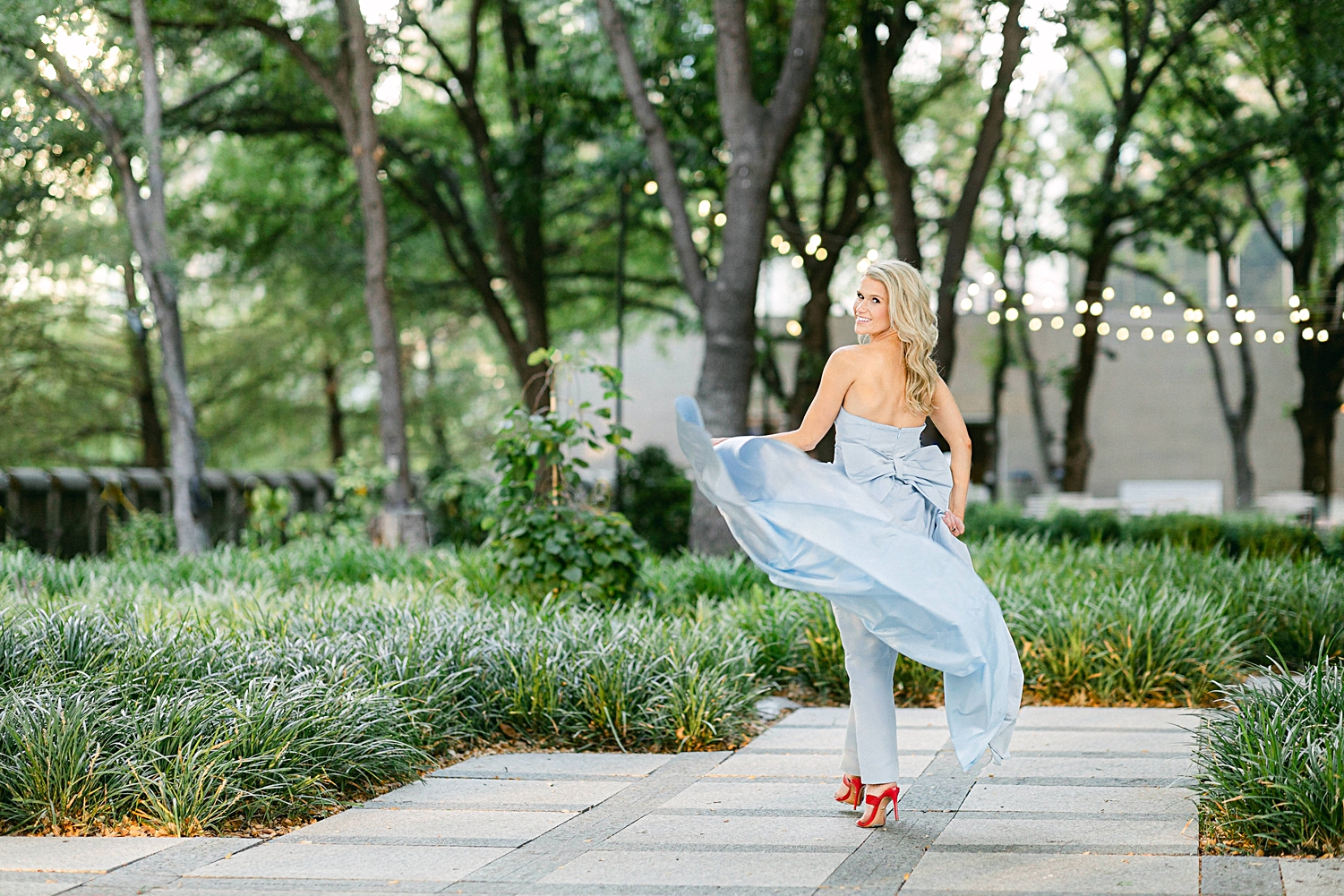 blonde woman laughing in light blue jumpsuit with skirt and big bow on back land red shoes in green urban garden