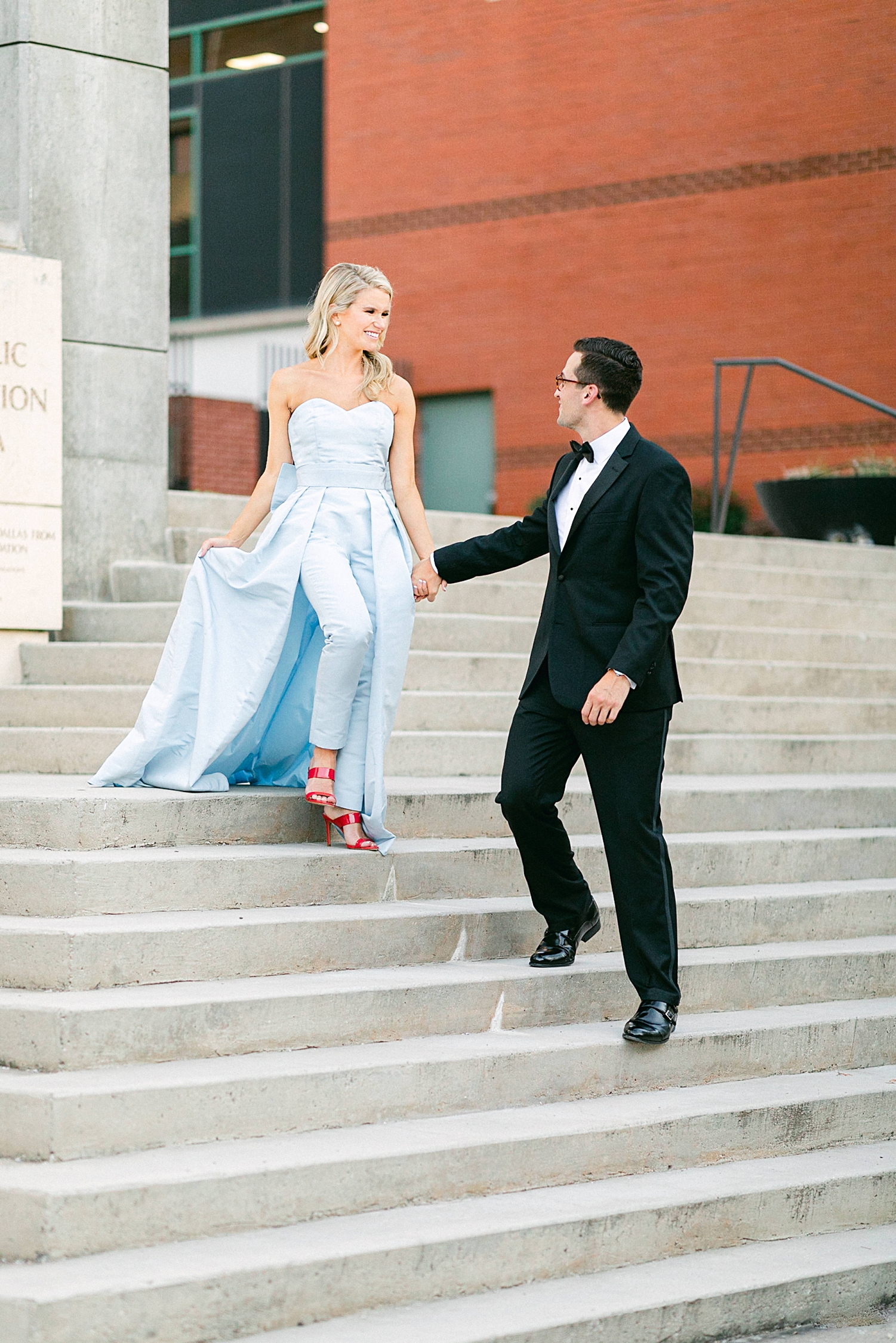 Man in black tuxedo and woman in light blue jumpsuit walking down stairs in downtown Dallas engagement
