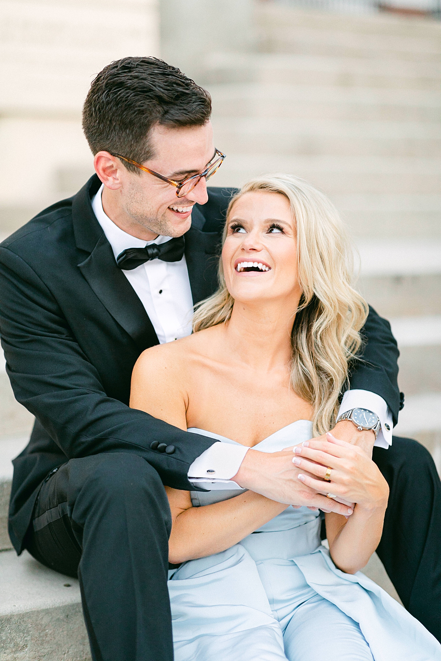 Man in black tuxedo and woman in light blue jumpsuit sitting on stairs in downtown Dallas engagement