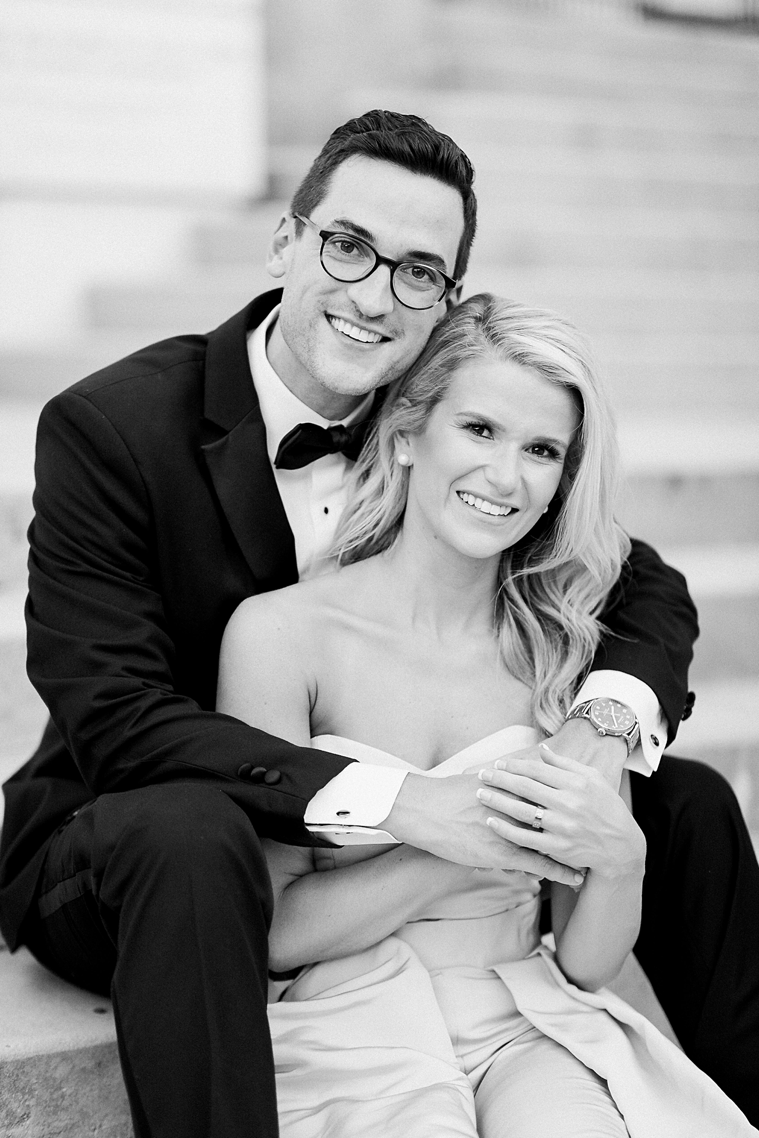 Man in black tuxedo with arms around woman in light blue jumpsuit sitting on stairs smiling