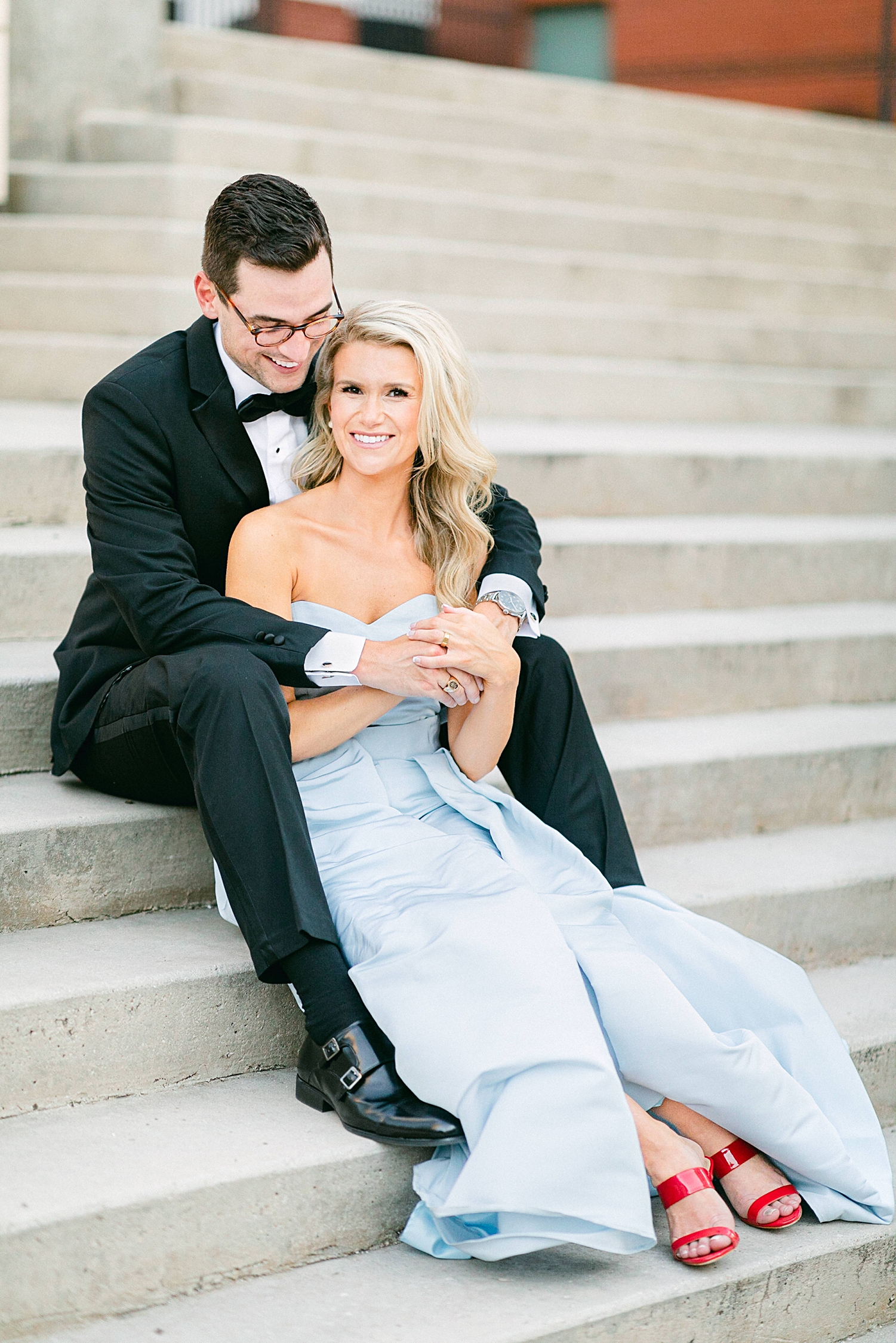 Man in black tuxedo with arms around woman in light blue jumpsuit and red shoes sitting on stairs in downtown Dallas engagement session