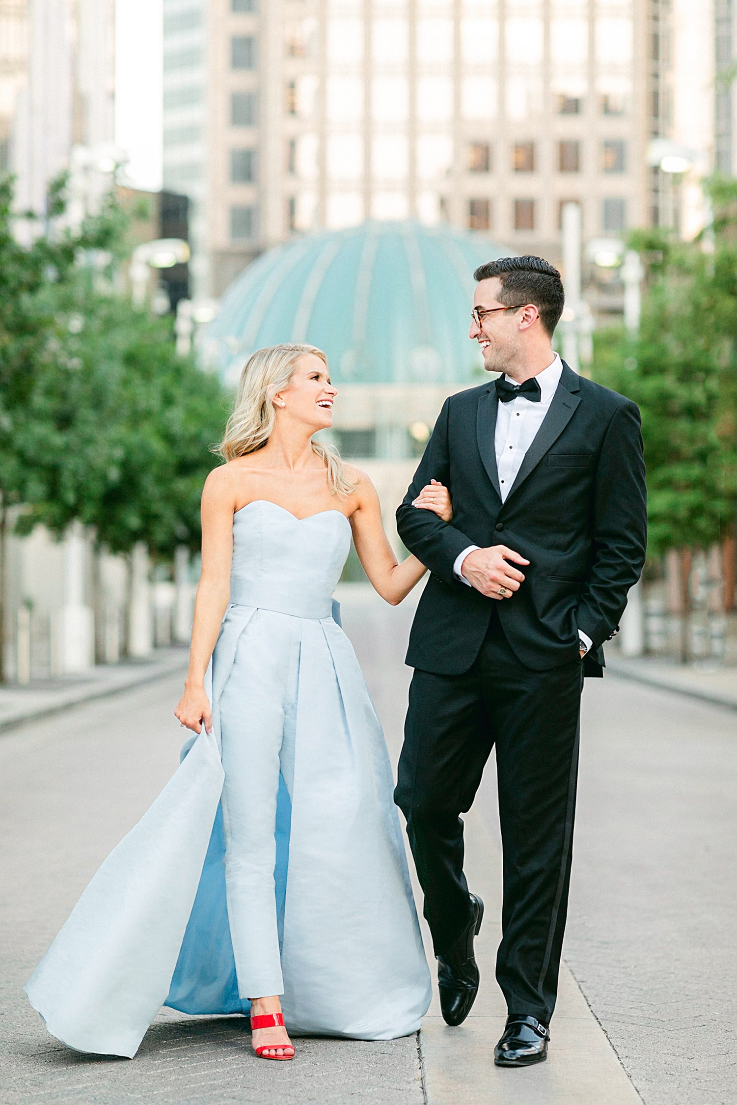 Man in black tuxedo and woman in light blue jumpsuit and red shoes walking side by side in downtown Dallas engagement session