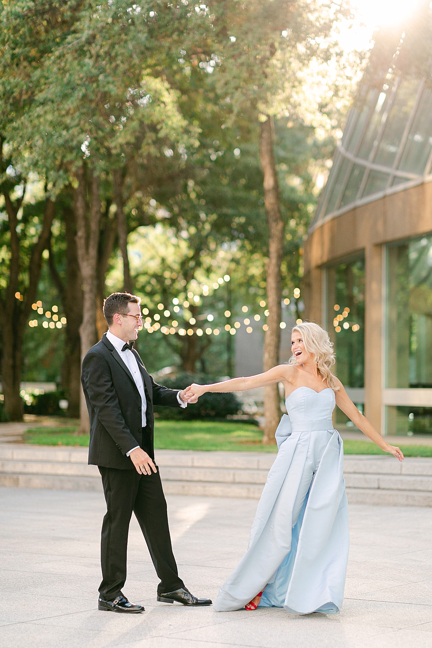 Man in black tuxedo and woman in light blue jumpsuit dancing by green garden in downtown Dallas engagement