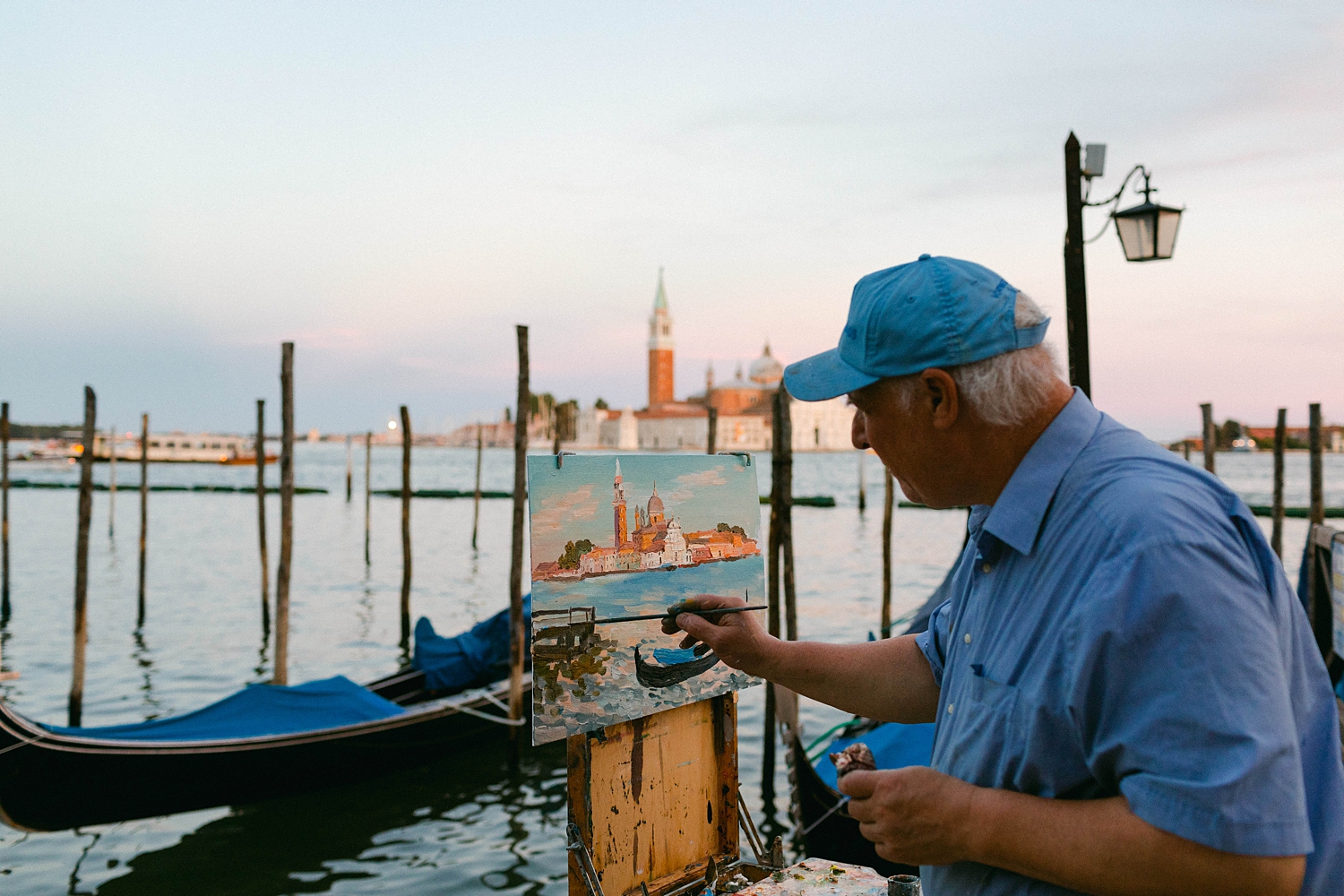man painting image of Grand canal in Venice church tower and blue gondolas against sunset