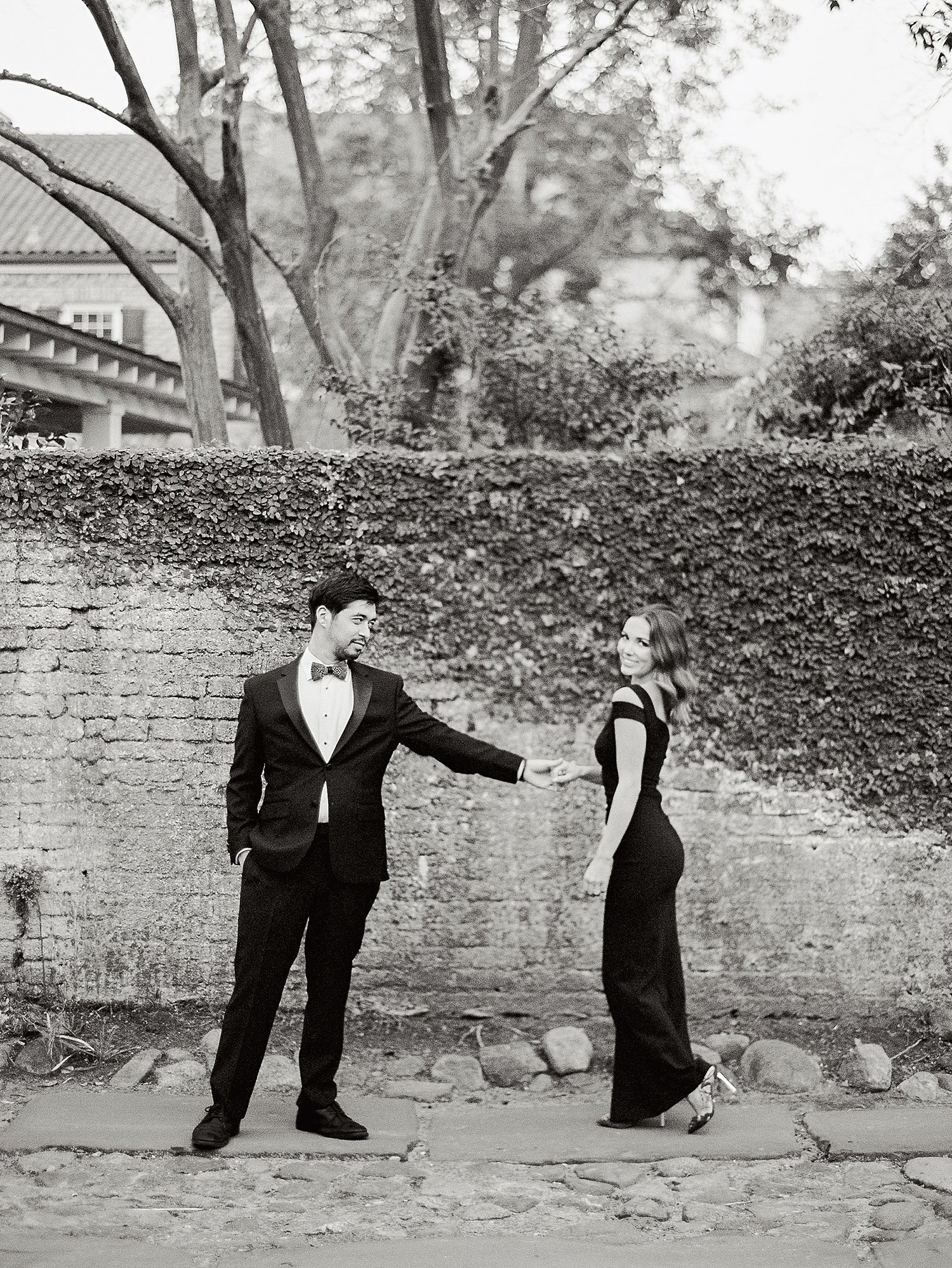 Man in tuxedo reaching hand out for woman in black dress on cobblestone alley and brick wall